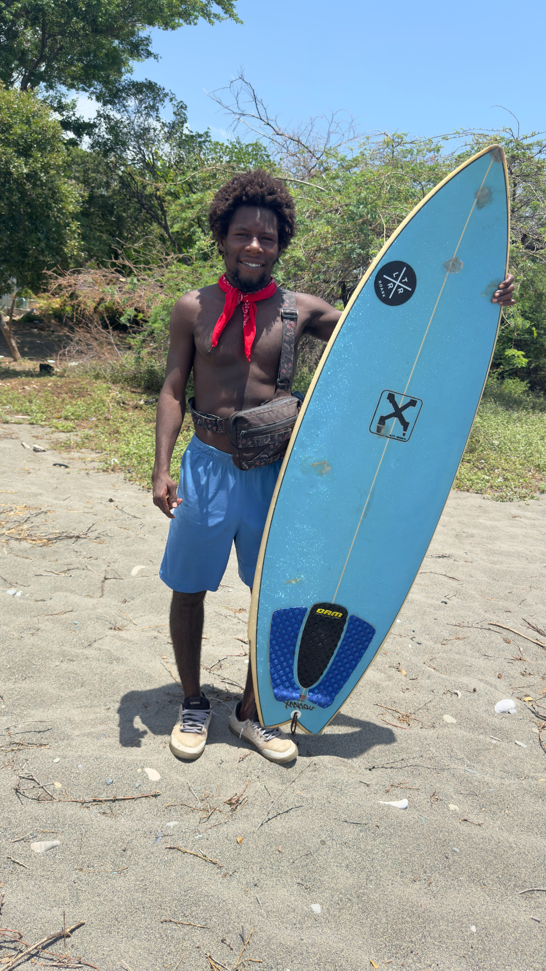 A shirtless man with a red bandana around his neck holding a surfboard at Jamnesia surf camp on the beach, with trees and sand in the background.