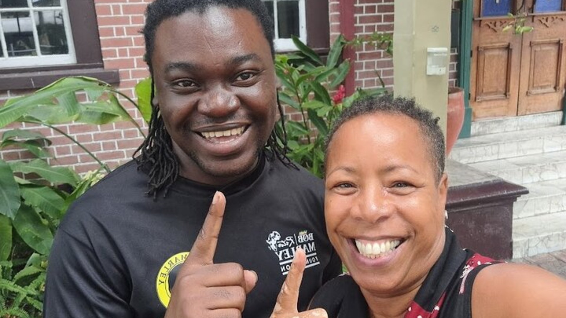 A man and woman smiling and taking a selfie outdoors in front of a brick building with plants around. Kgn Jamaica