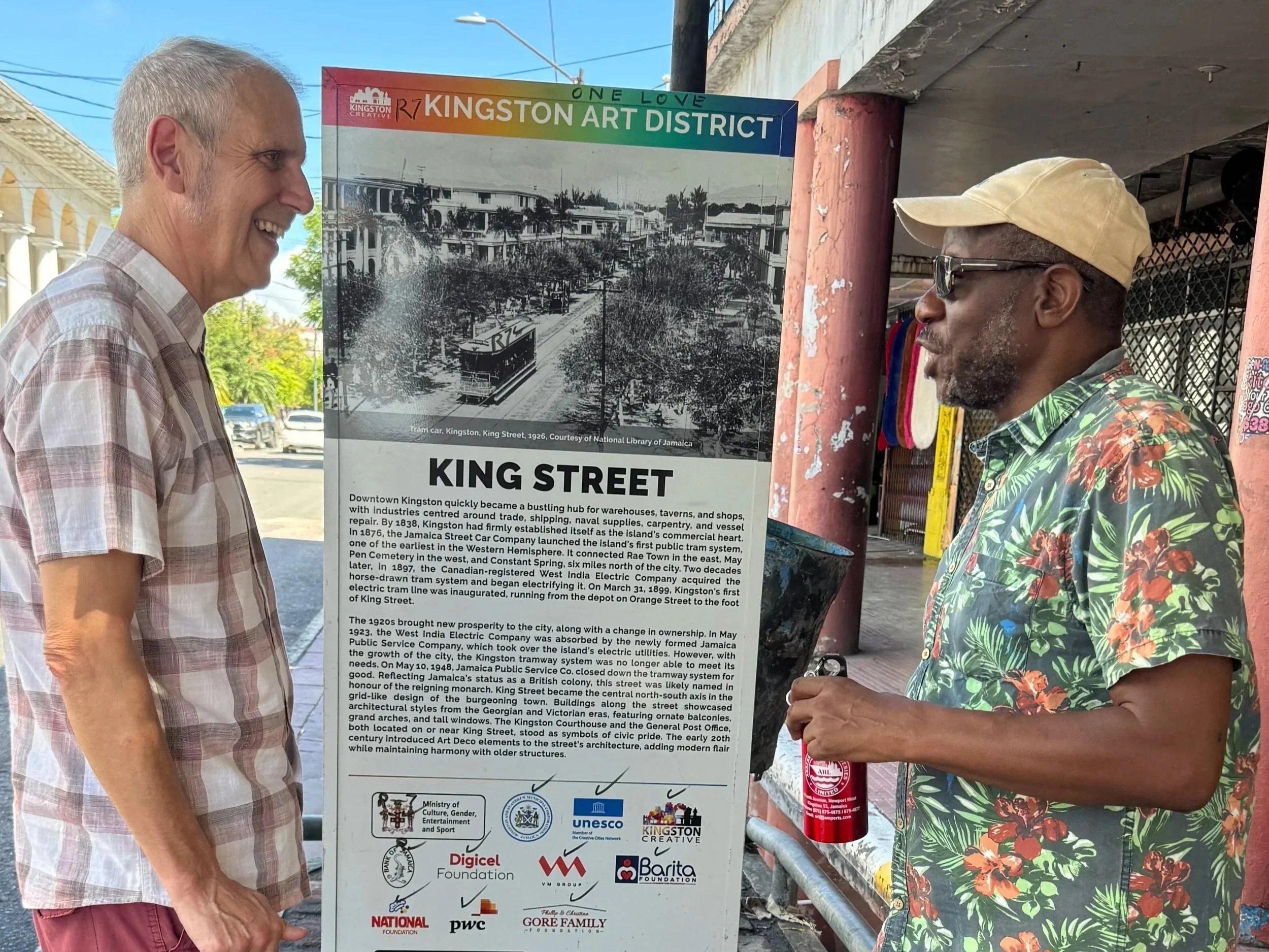 Two men stand on a sidewalk reading an informational sign about Kingston's history. One man wears a plaid shirt and the other a floral Hawaiian shirt with a cap. The sign features historical photos and logos of organizations.
