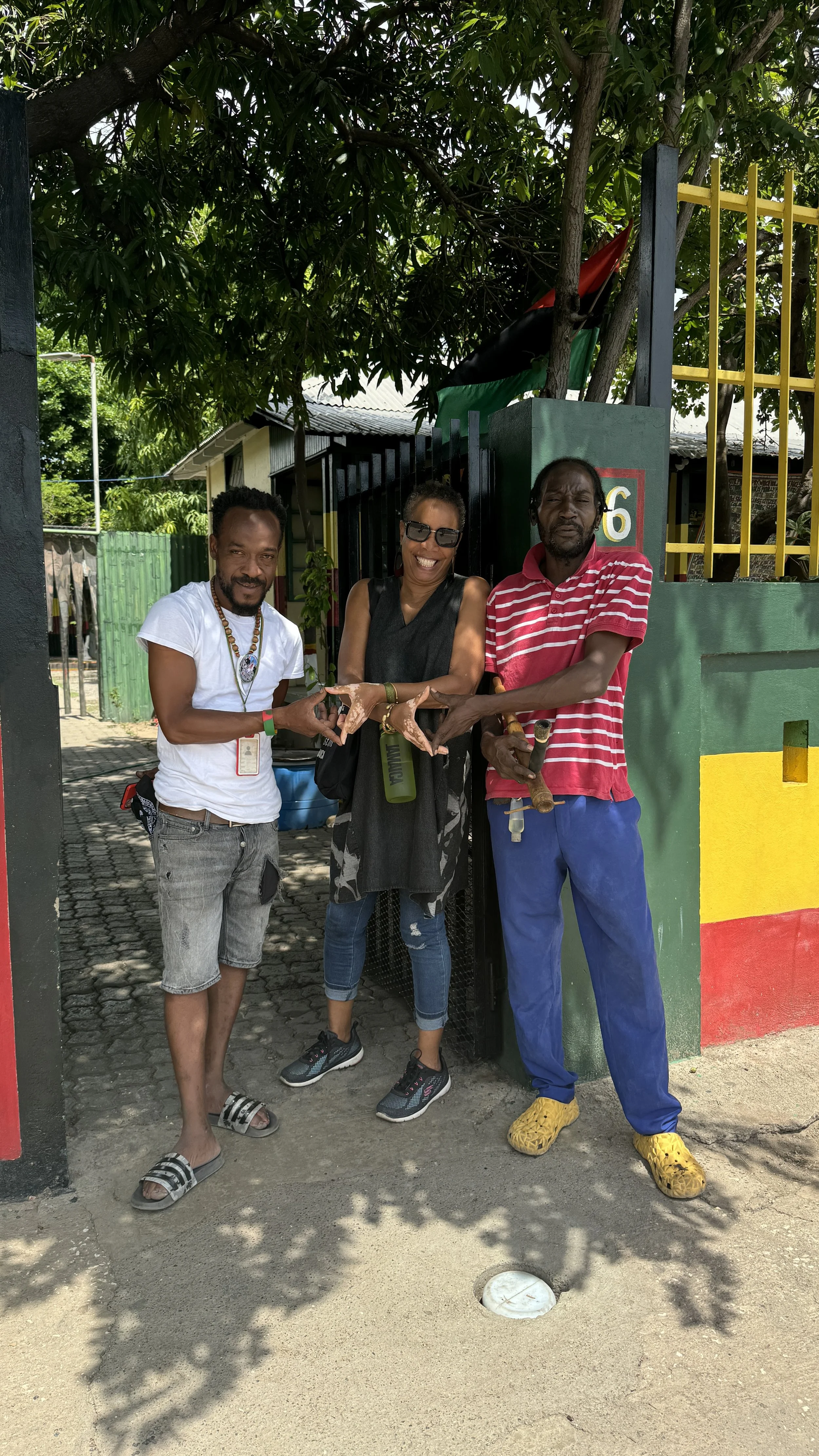 Three people standing together outdoors near a fence, with foliage overhead. The person on the left is a man wearing a white t-shirt, gray shorts, and slide sandals. The person in the middle is a woman wearing sunglasses, a black sleeveless top, and ripped jeans. The person on the right is a man wearing a red and white striped shirt, blue pants, and yellow shoes. They are all smiling and making hand gestures.