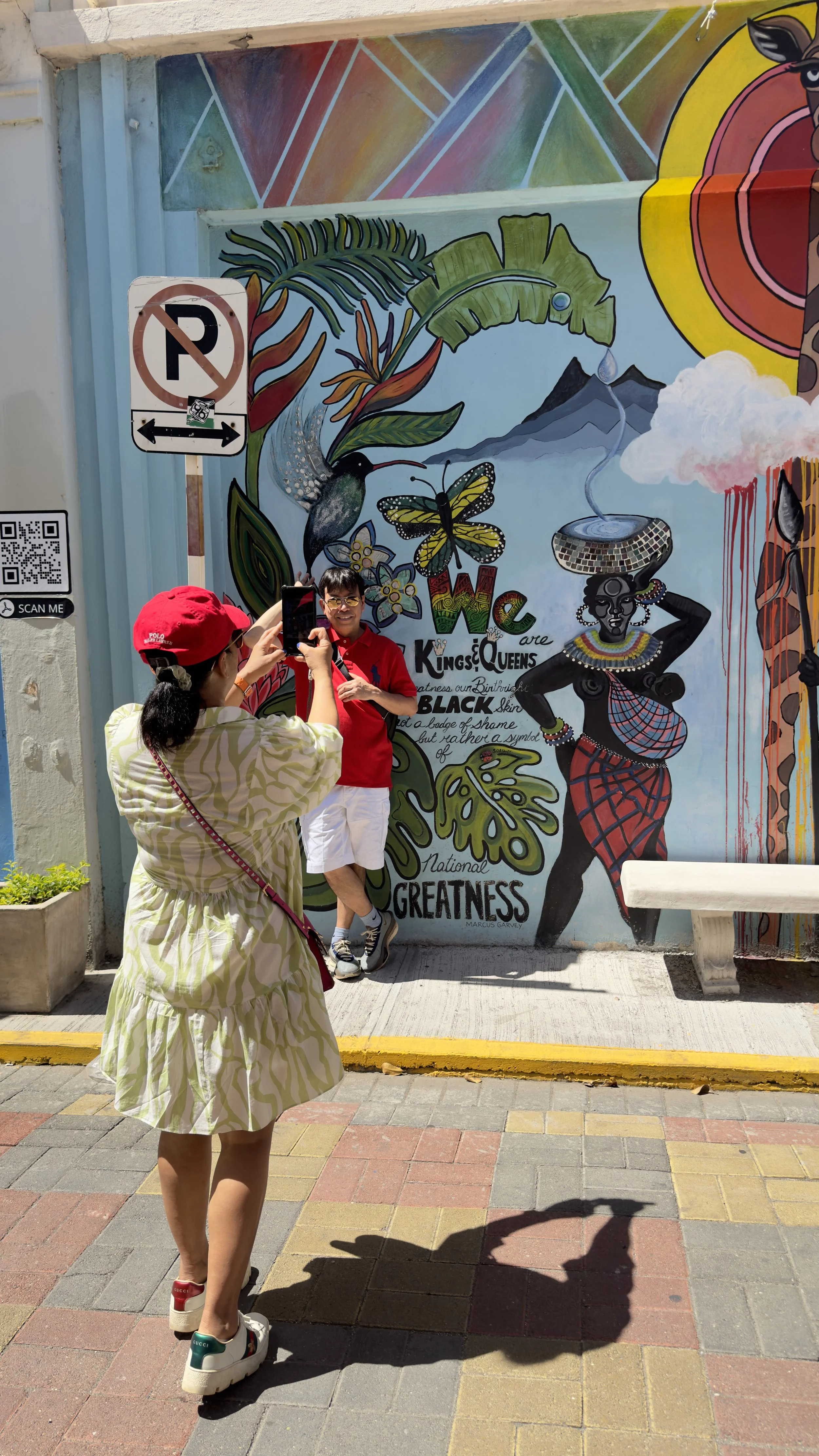 A woman taking a picture of a man standing against a colorful mural. The mural features various vibrant illustrations including a woman in traditional attire, a hummingbird, butterfly, and lush leaves with the words 'We are Kings & Queens' and 'Great