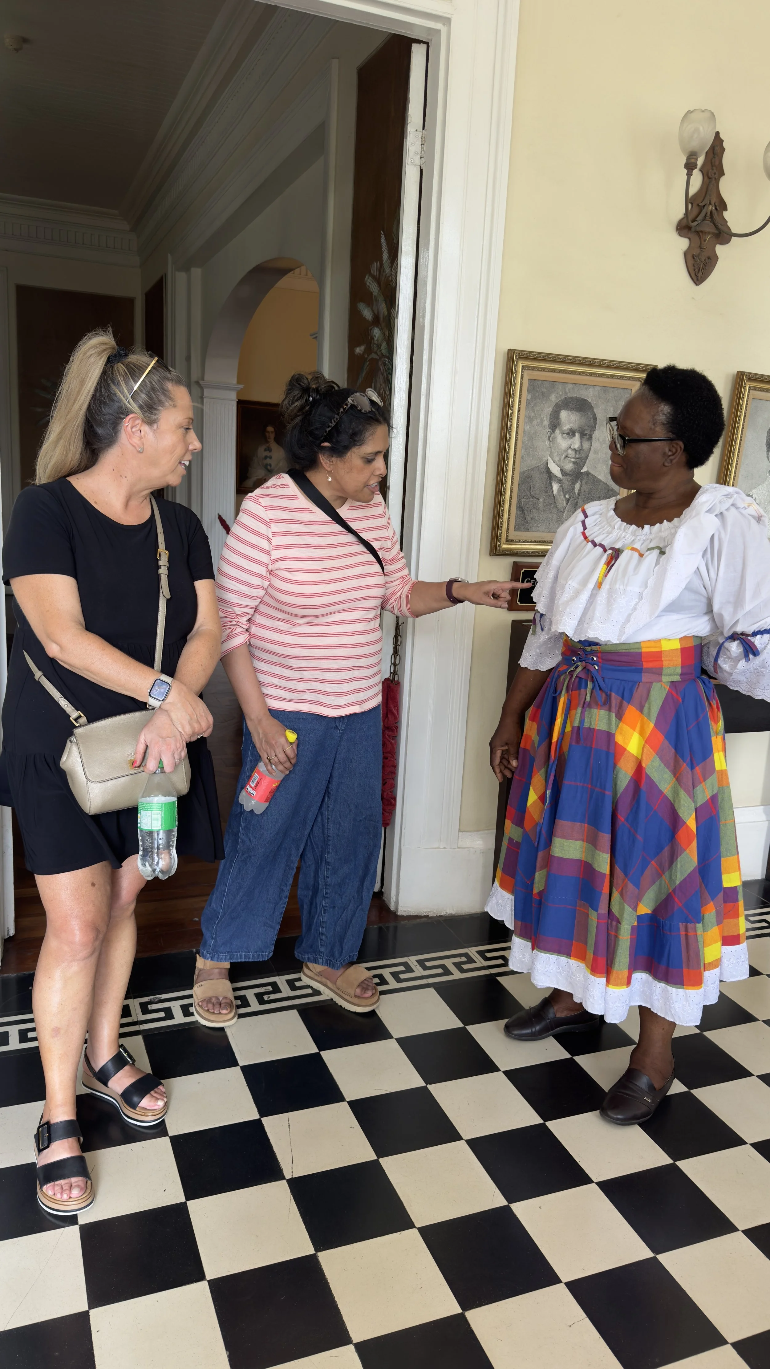Three women of different ethnicities in conversation inside a historic house, with black and white floor and framed black and white portraits on the wall. Located at one of Kingston's top attractions Devon house.