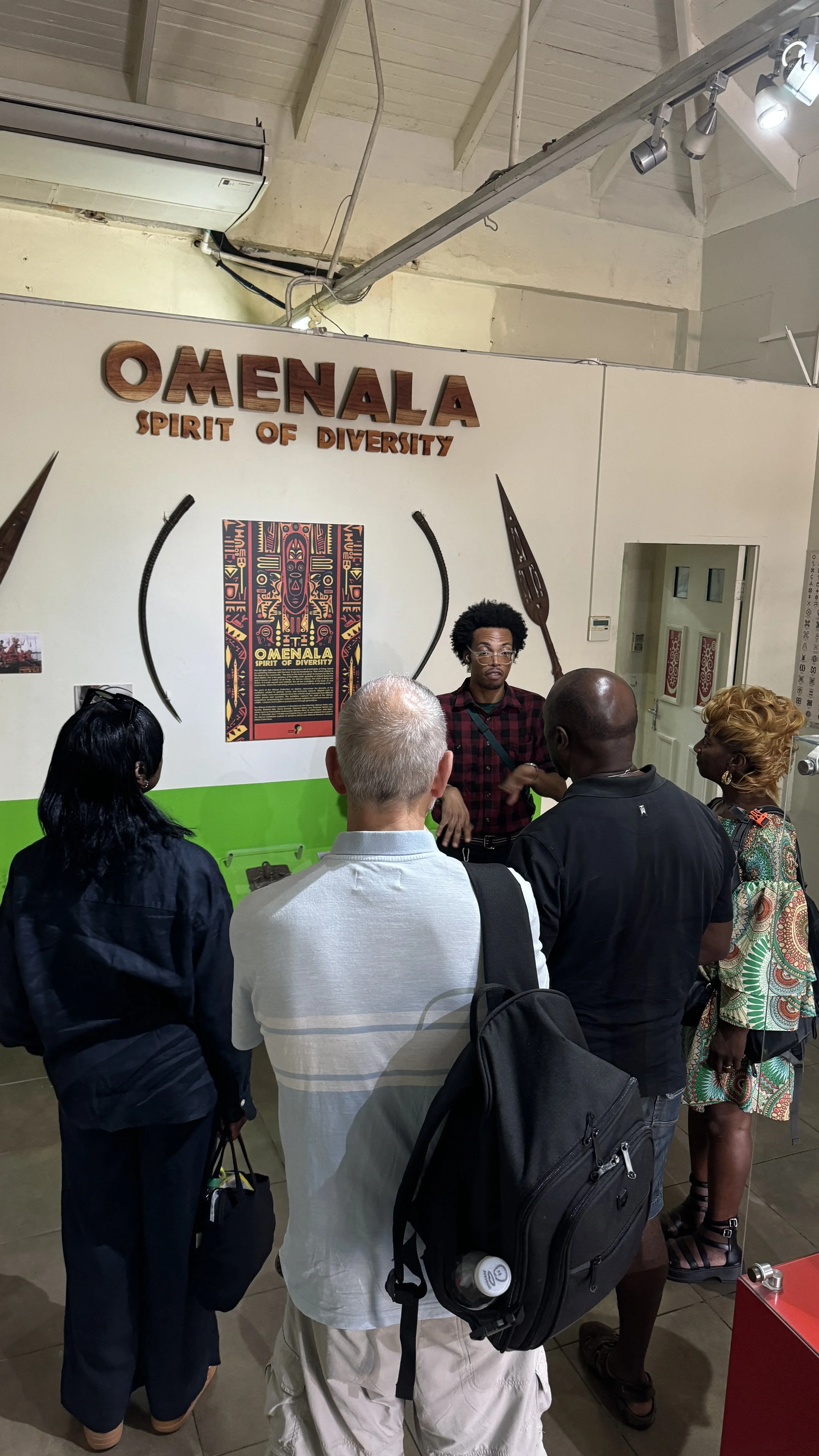 Group of people listening to a guide in front of a display about Omenala, the spirit of diversity, with traditional Tongan spears and a colorful poster.