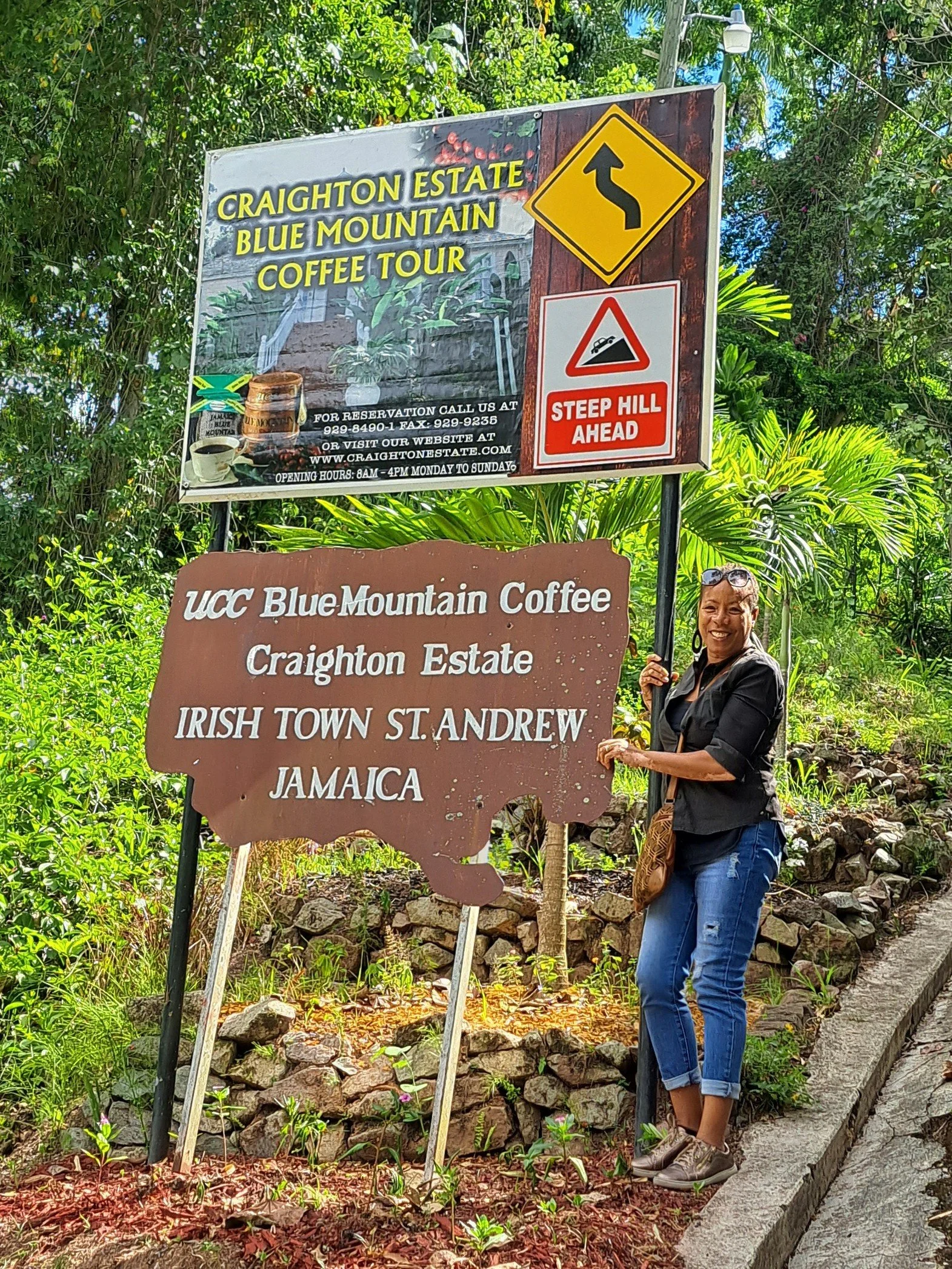 A woman standing next to a brown sign and smiling; the sign reads: "UCC Blue Mountain Coffee, Craighton Estate, Irish Town St. Andrew, Jamaica". Above her are two other signs: one yellow with a curvy arrow indicating a winding road, and another white and red warning sign stating "Steep Hill Ahead". There are lush green plants and trees in the background.