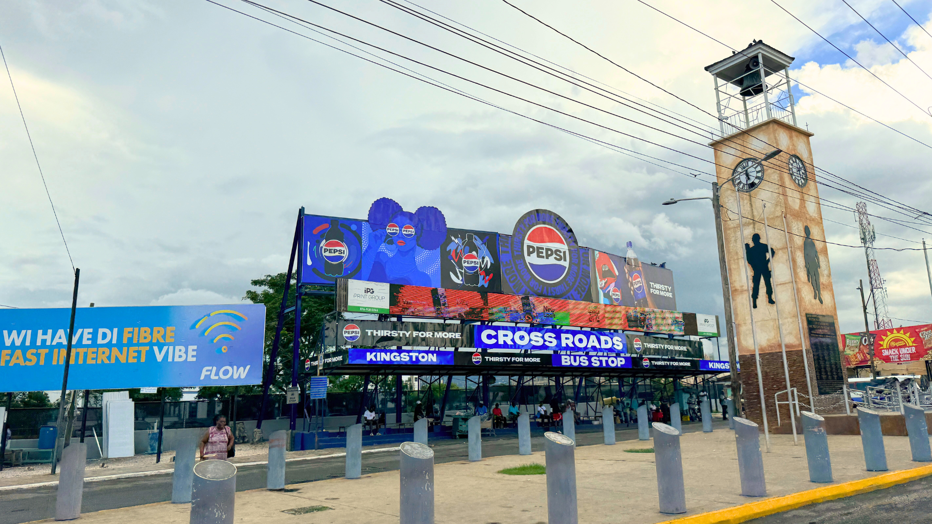 A bus stop with a large electronic billboard displaying Pepsi advertisements, located near a tall clock tower with silhouettes of a man and a woman painted on it, and a blue sign on the left promoting fiber internet. This is located on Cross roads Ki