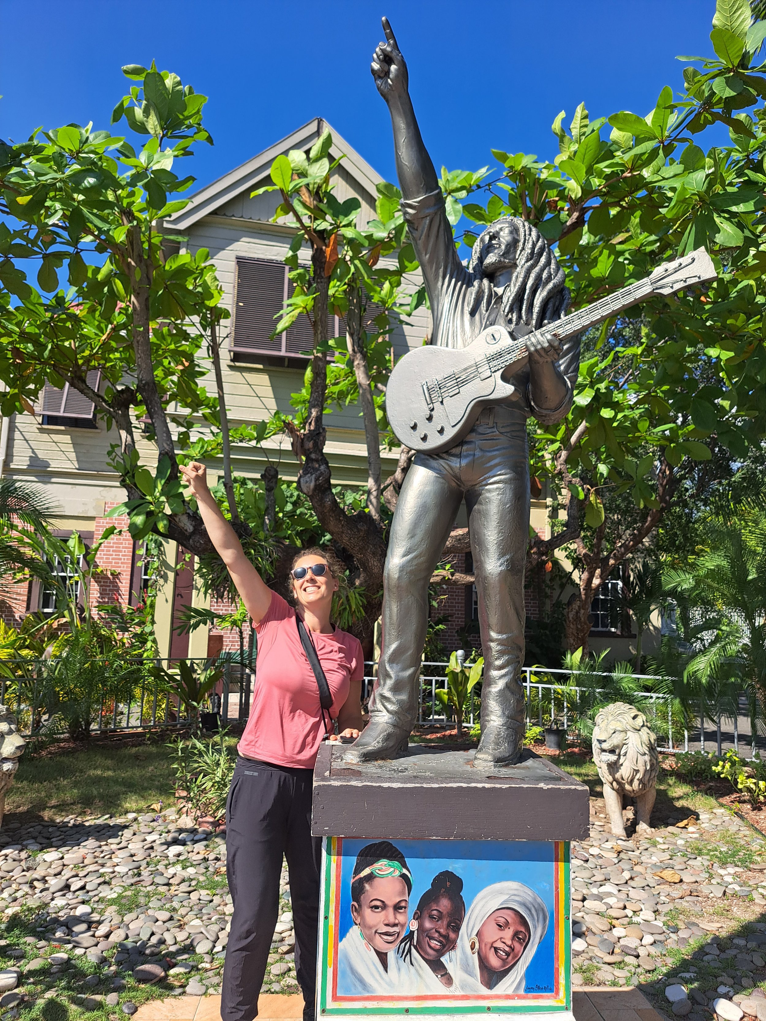 A woman with sunglasses, a pink t-shirt, and dark pants standing next to a large statue of Bob Marley playing guitar with one had raised to the sky. While on tour in Kingston, Jamaica with Onestop cultural tours Jamaica.