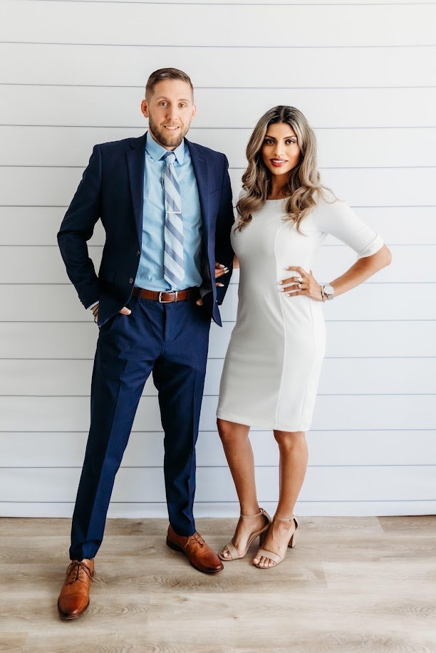 A man and woman standing side by side against a white wooden wall, dressed professionally. The man is in a navy suit with a light blue shirt and striped tie, and the woman is in a white dress with beige heels.