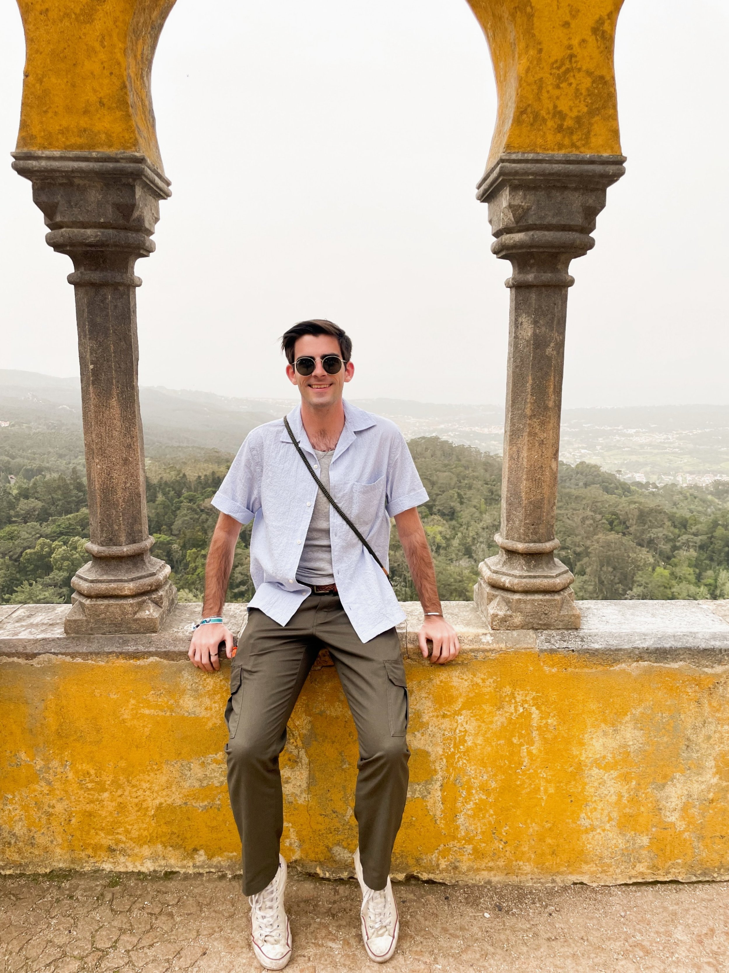 A smiling young man in sunglasses, a light blue short-sleeve shirt, and cargo pants, sitting on a brick ledge beneath a historical stone and yellow wall arch, with a scenic view of green hills and a cloudy sky in the background.