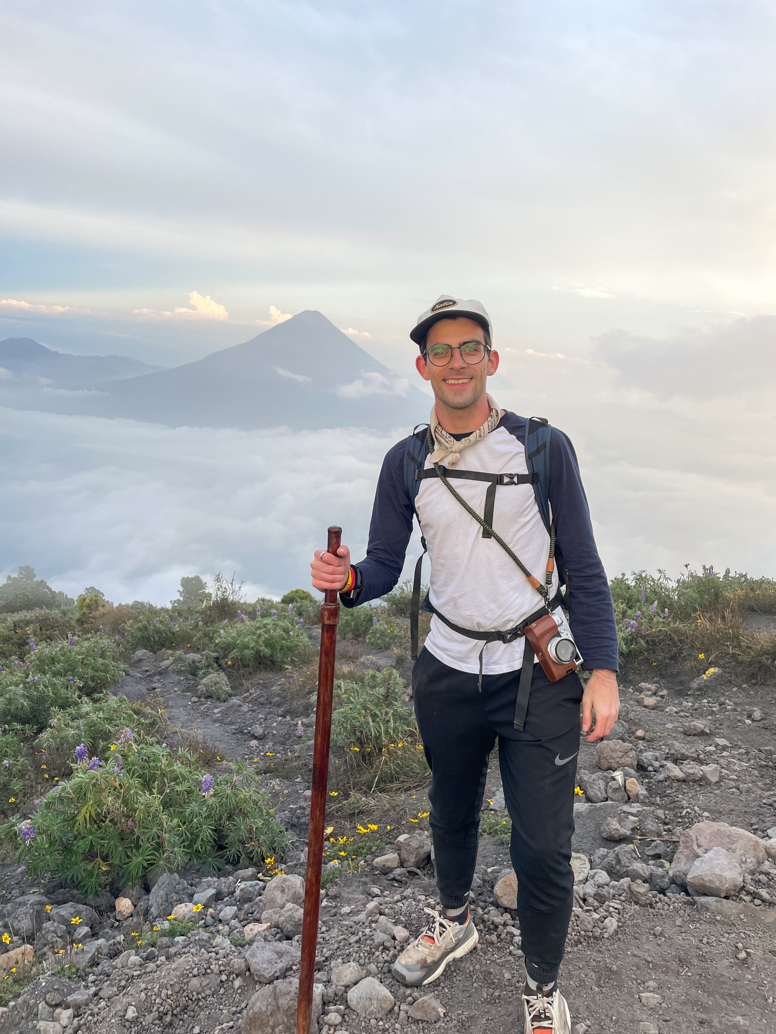 A man standing on a rocky mountain trail with a walking stick, smiling, with a large volcano and clouds in the background.