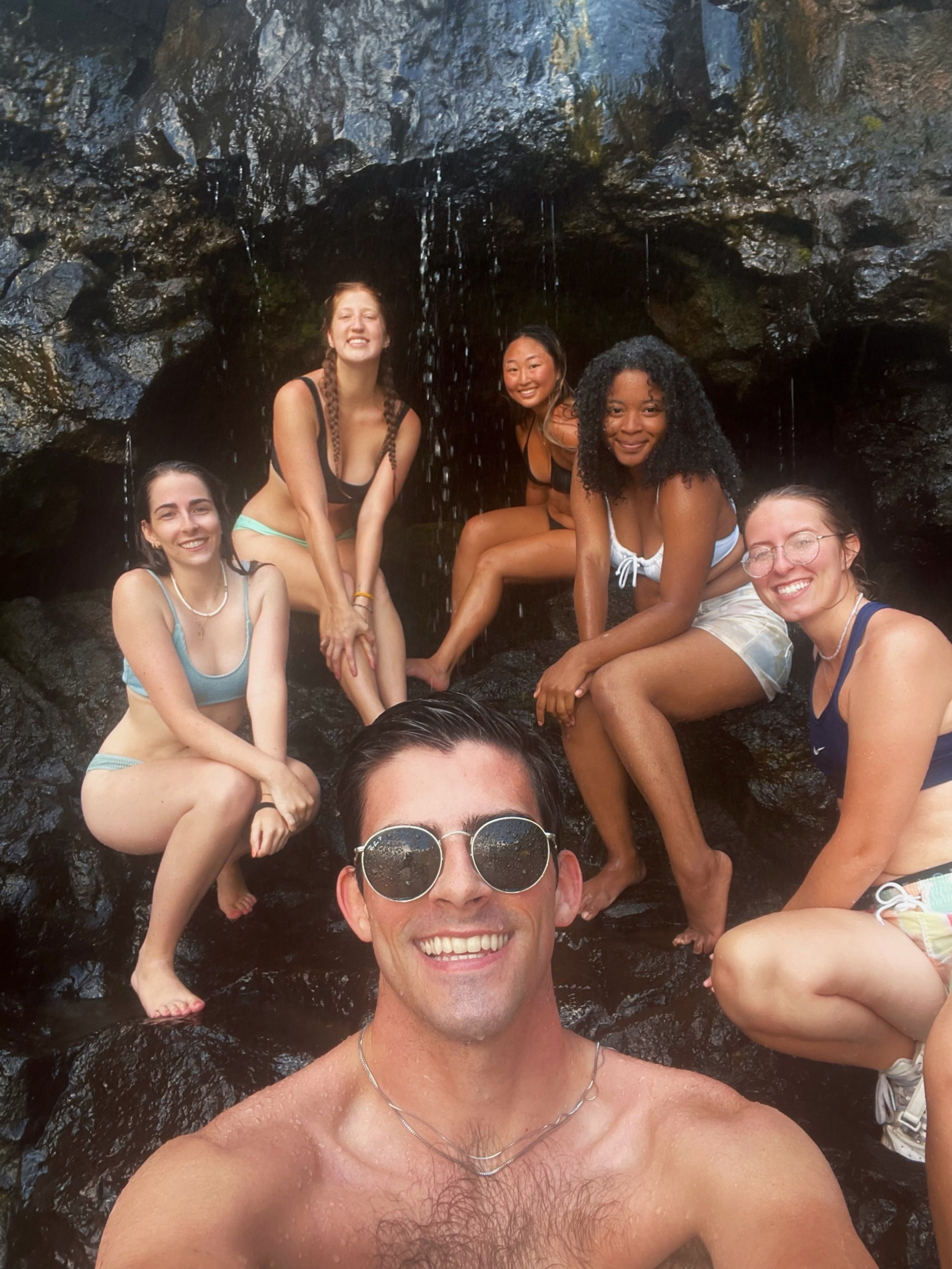 Group of six friends in swimsuits at a waterfall, smiling, with one person taking a selfie in the foreground.