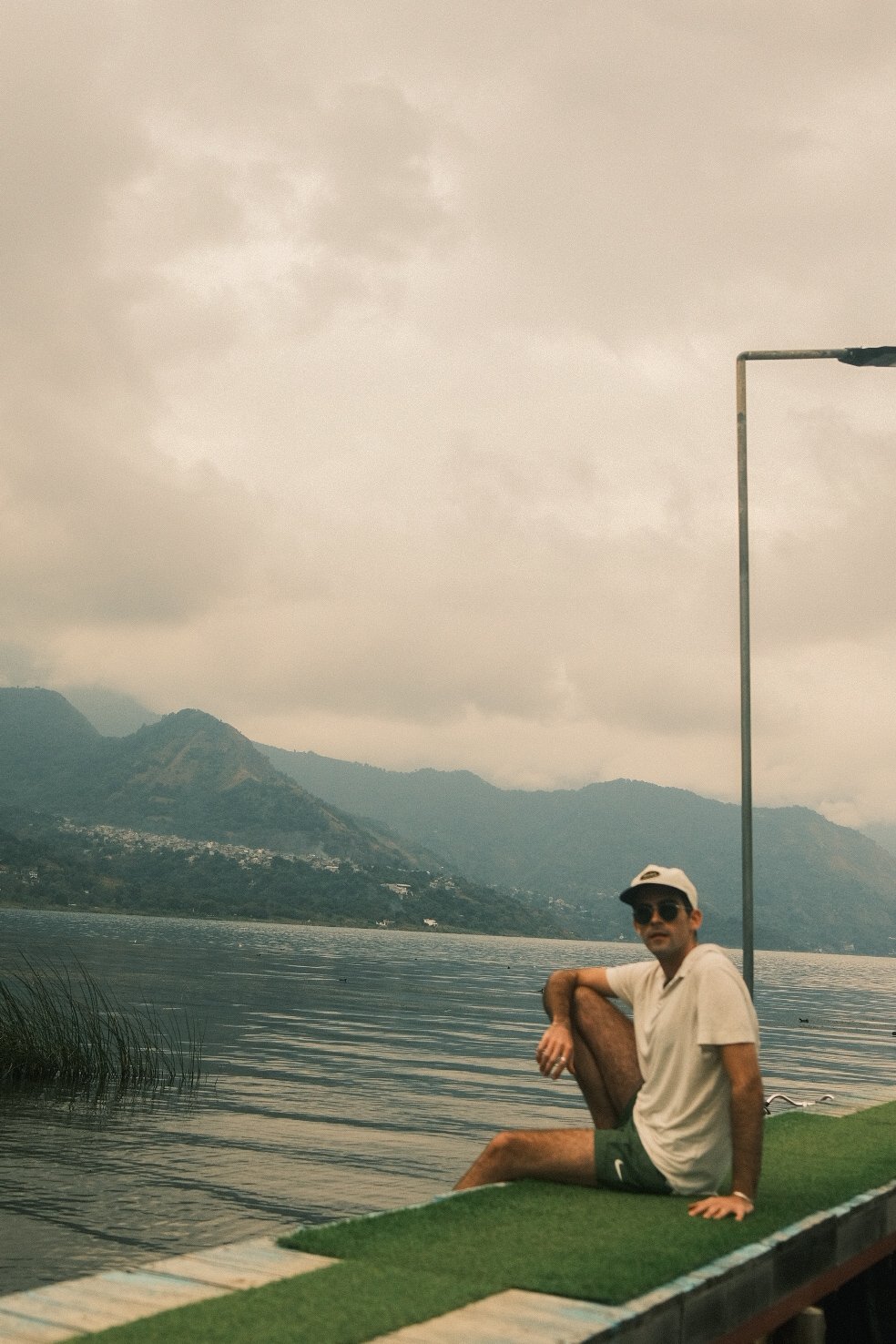 A man wearing sunglasses, a cap, a beige shirt, and shorts, sitting on a small platform covered with green artificial grass beside a calm lake with mountains in the background and cloudy sky above.