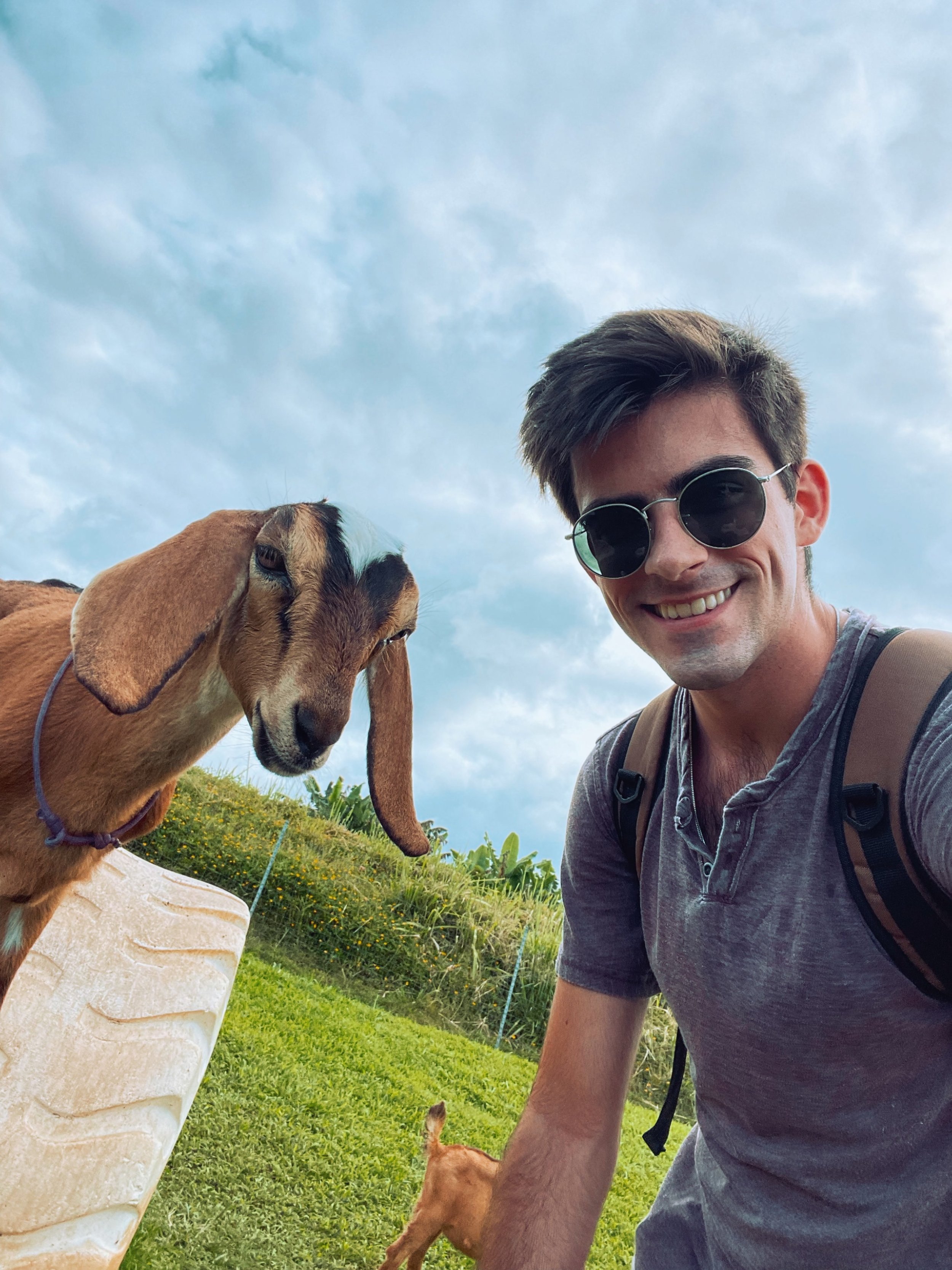 A young man smiling for a selfie outdoors next to a goat with long ears, under a partly cloudy sky.