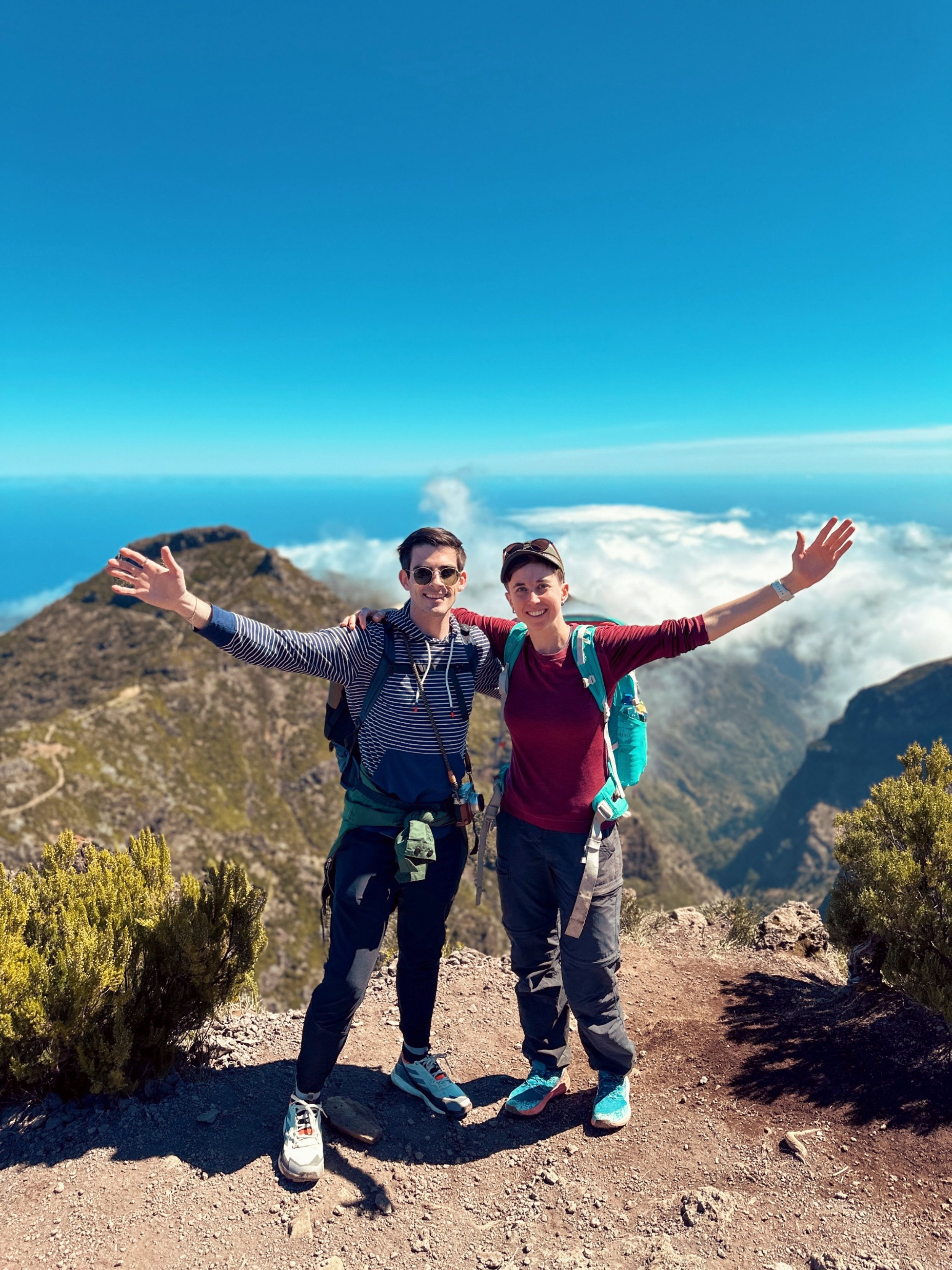 Two hikers with backpacks and outdoor clothing standing on a mountain trail with arms outstretched, smiling, with a mountain landscape, blue sky, and clouds in the background.
