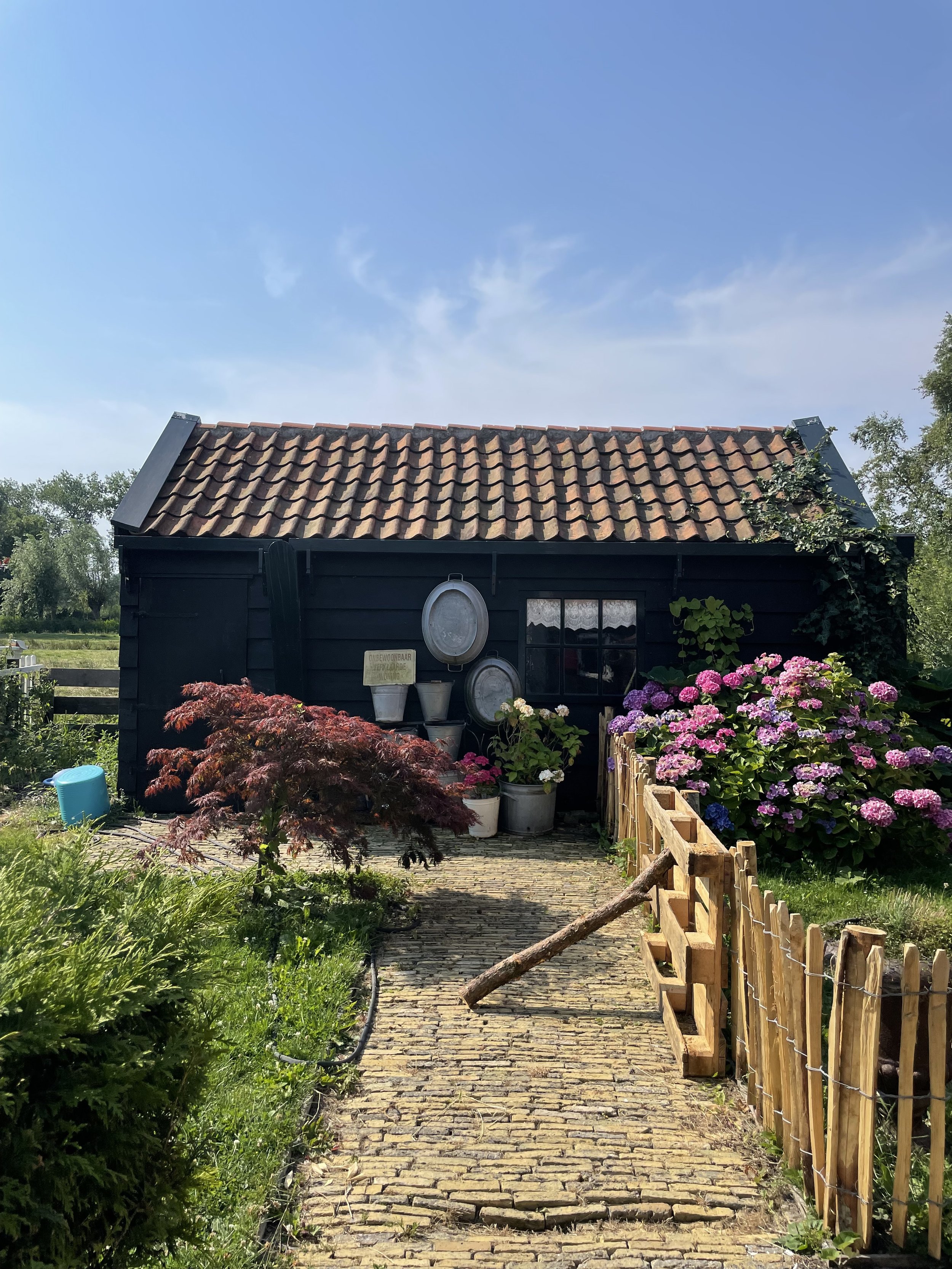 A small black wooden shed with a terracotta tiled roof, surrounded by colorful flowers and a cobblestone pathway, with a clear blue sky overhead.