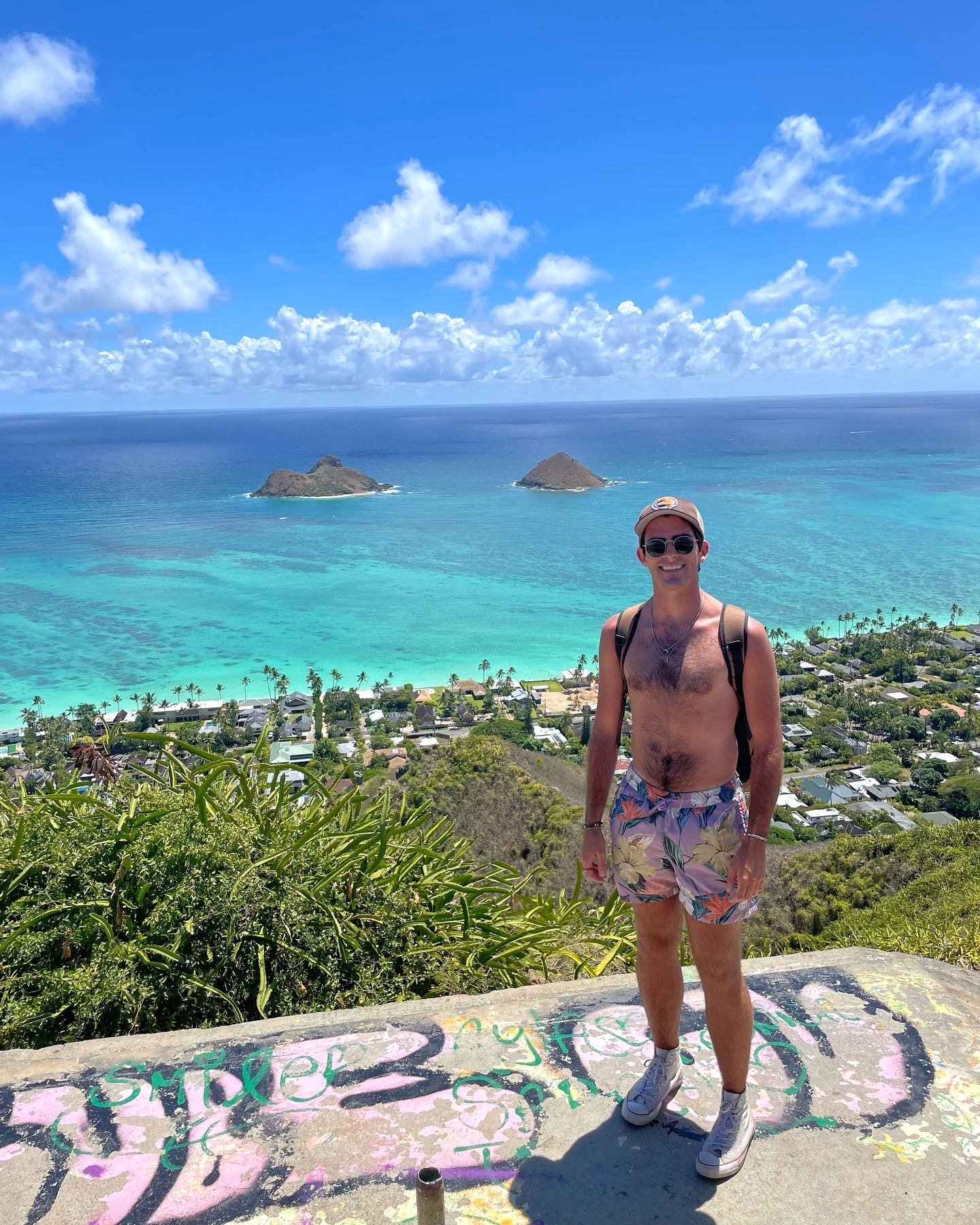 A young man with sunglasses, a backpack, and floral shorts standing on a hilltop with a view of a tropical ocean, small islands, green vegetation, and a blue sky with clouds.