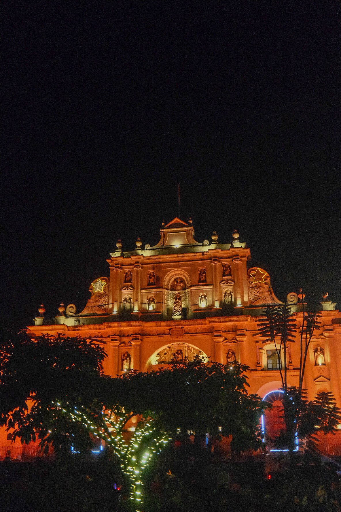 Night view of a well-lit baroque-style church decorated with stars and Christmas lights, partially obscured by trees.