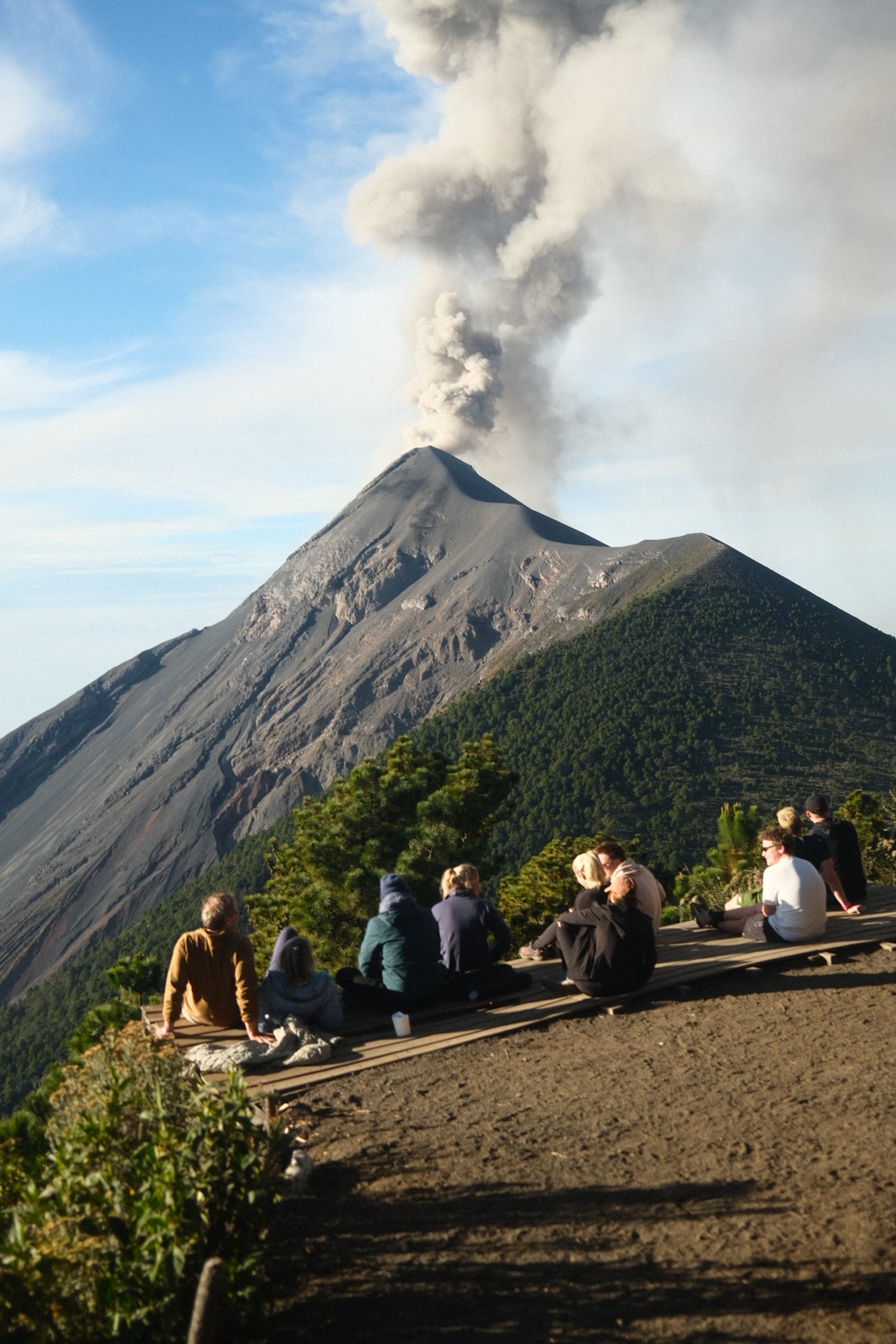 People sitting on a wooden platform observing erupting volcano with dark ash plume