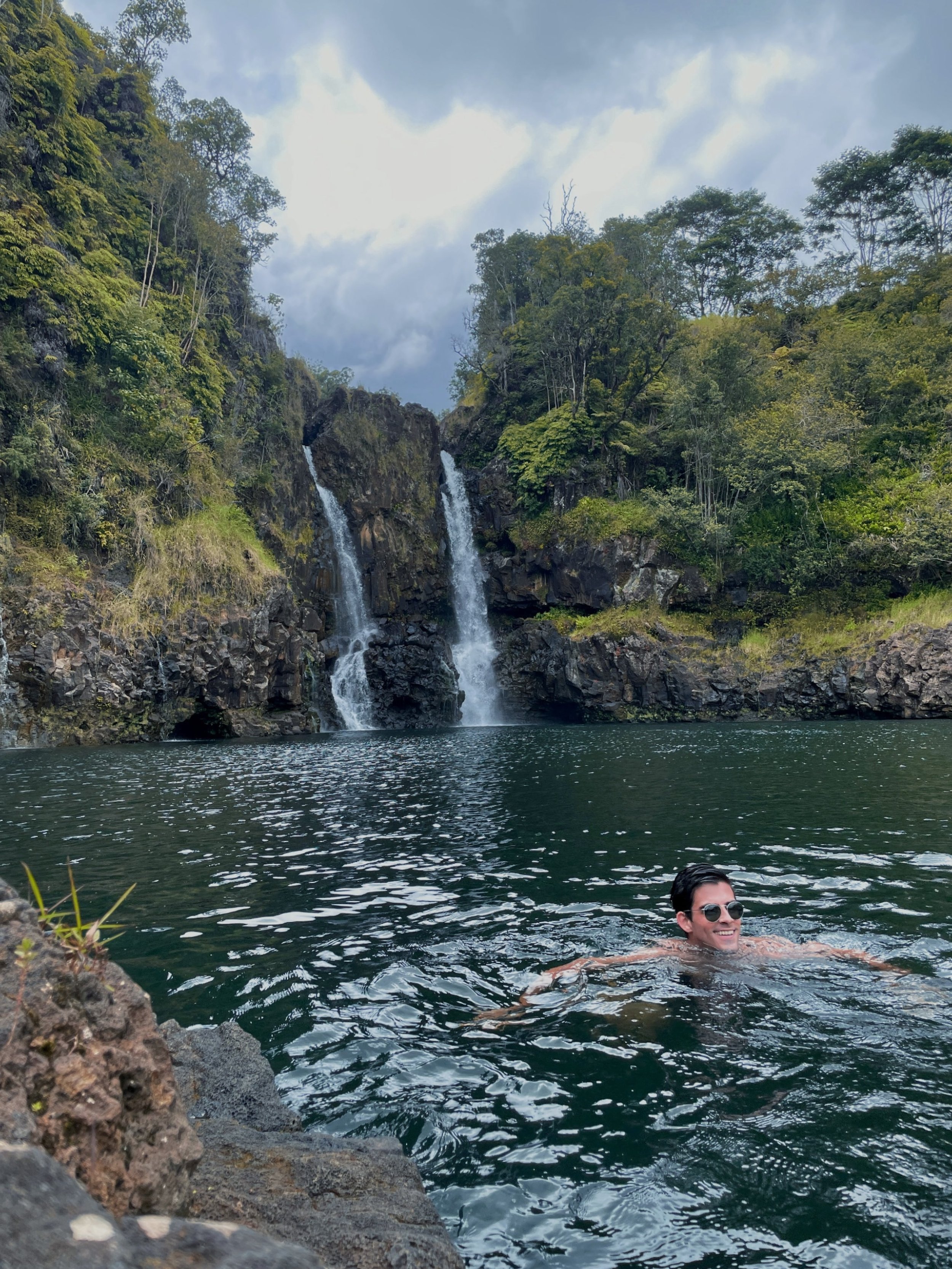 A man swimming in a natural body of water with a waterfall and lush greenery in the background.