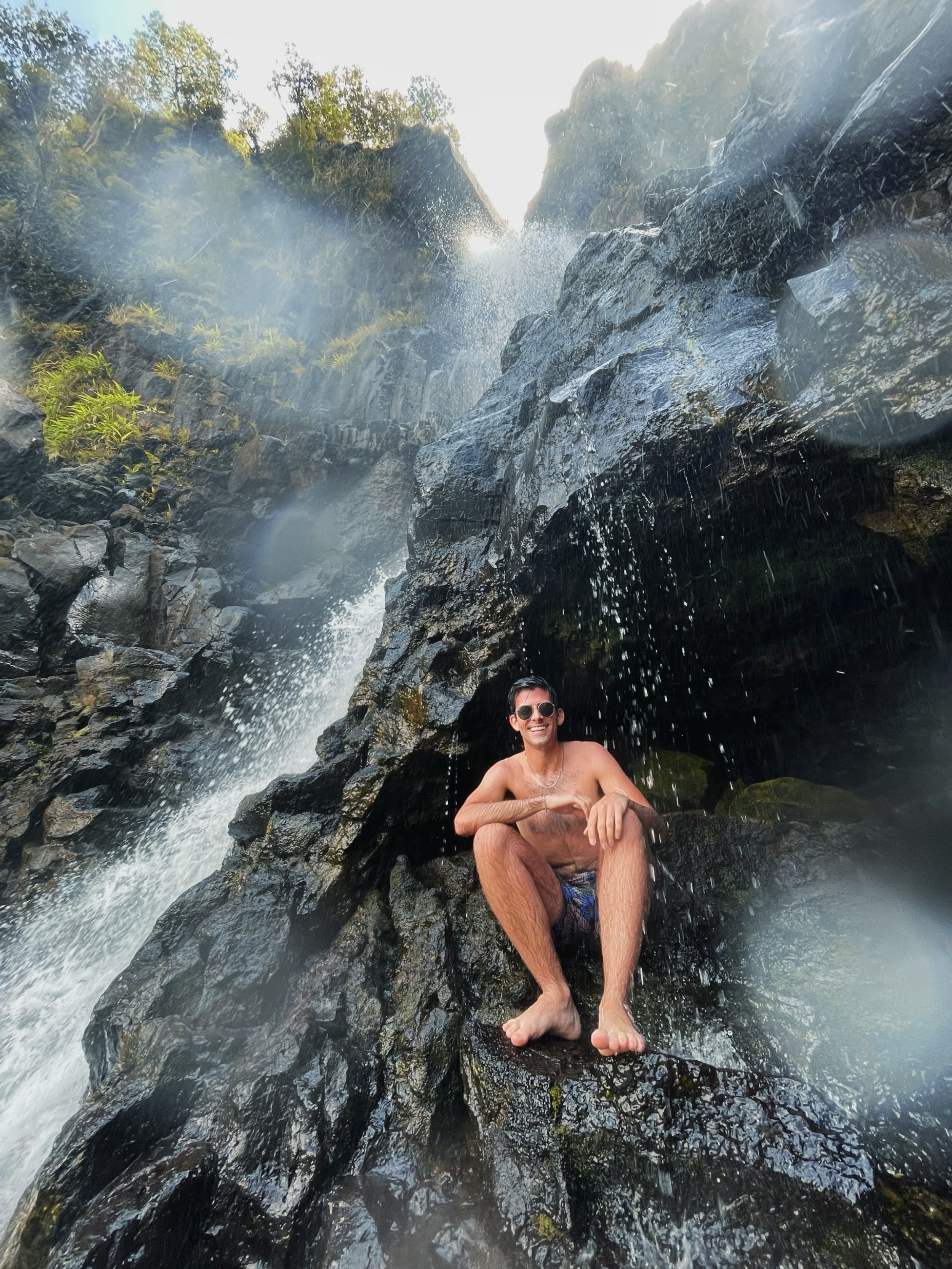 A man sitting on a wet rock under a small waterfall in a natural outdoor setting, wearing sunglasses and shorts, smiling at the camera.