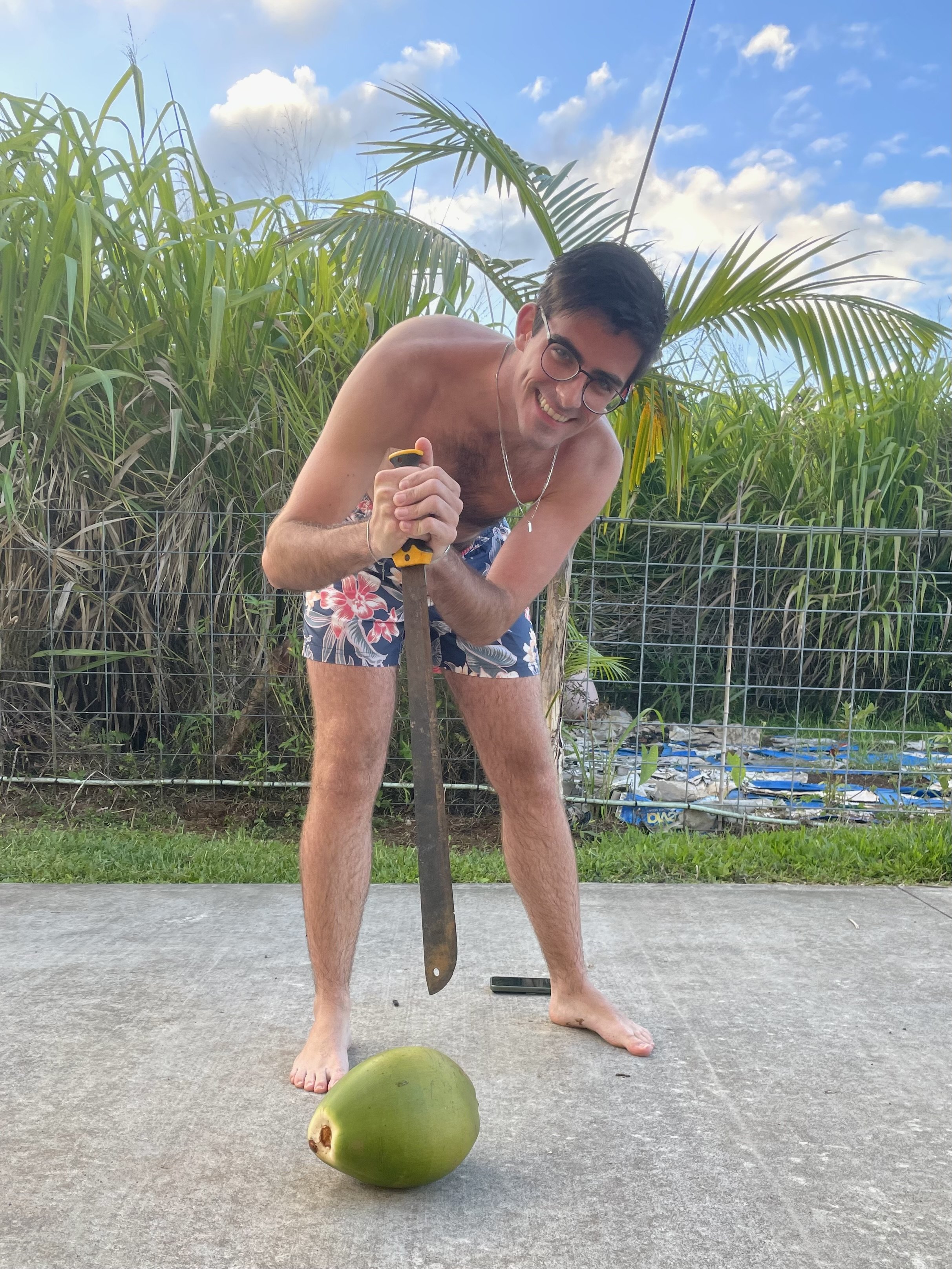 A young man with glasses, shirtless, wearing floral shorts, holding a large kitchen knife and smiling at the camera. He is standing on a concrete surface outdoors with a green coconut in front of him. The background shows tall green plants and a part