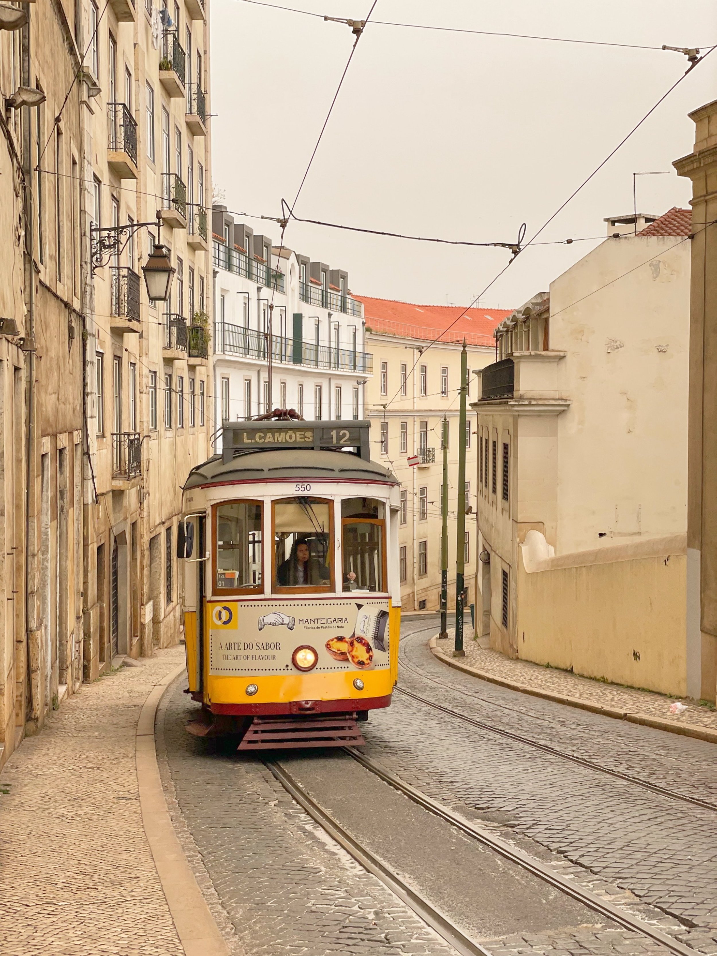 A yellow vintage tram traveling on cobblestone streets in an old European city, surrounded by tall buildings with balconies and street lamps.