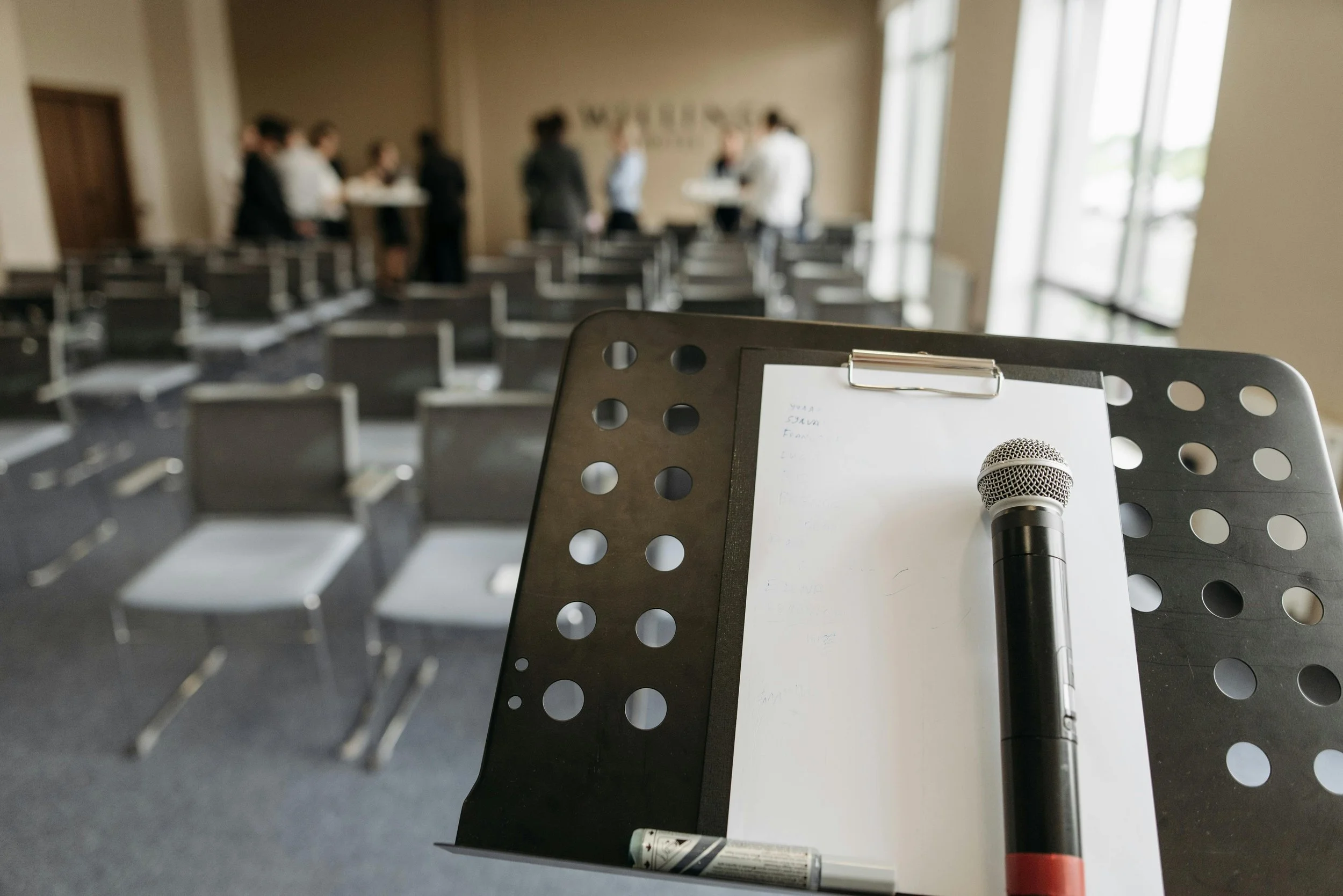 Public Speaking by Dr. DeJean, showing a microphone, paper, and marker on a music stand in the foreground, with a conference or seminar room filled with chairs and a group of people talking in the background.