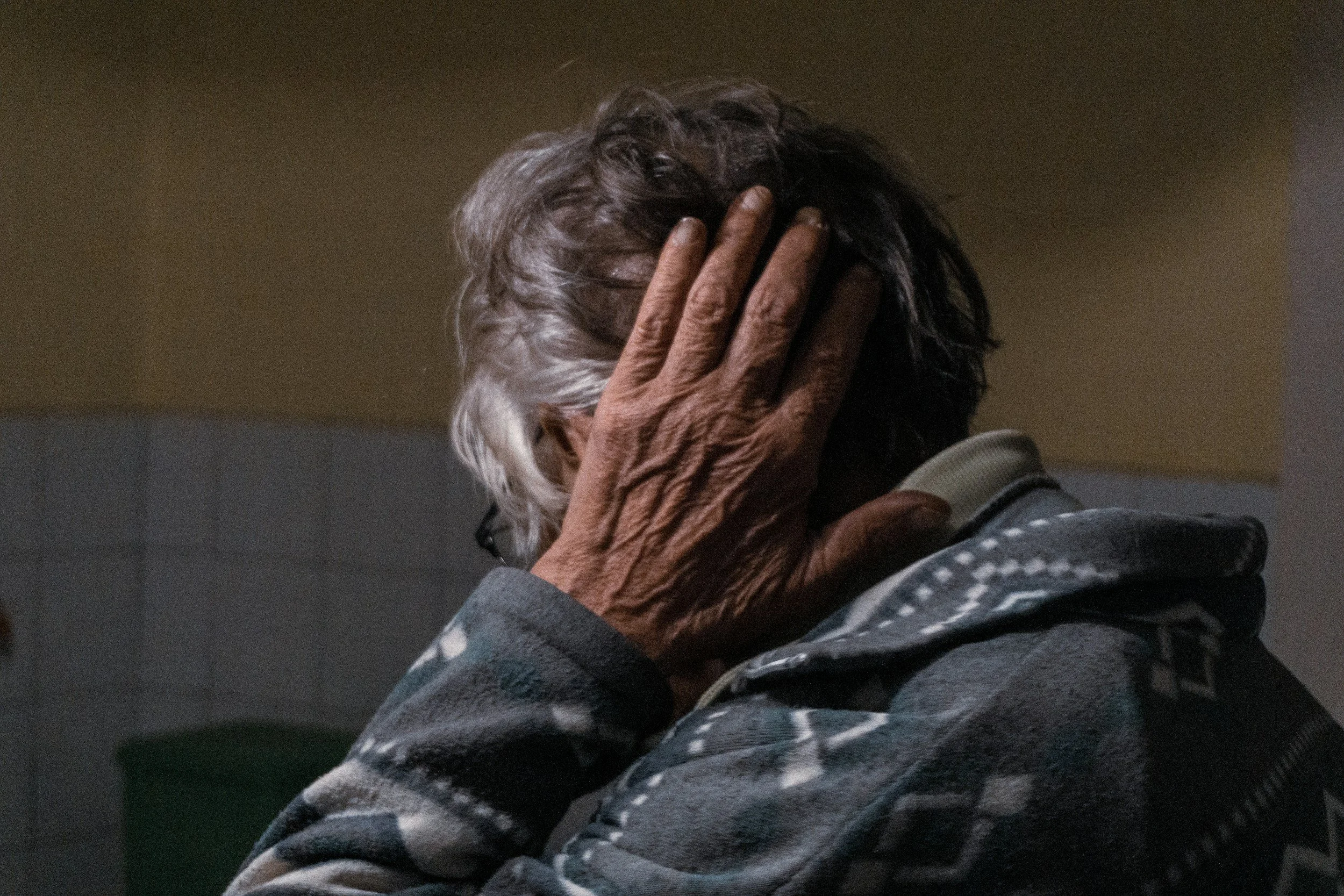 An elderly person with gray hair, wearing glasses and a patterned jacket, holds their head with one hand, covering their face in a gesture of distress or worry.