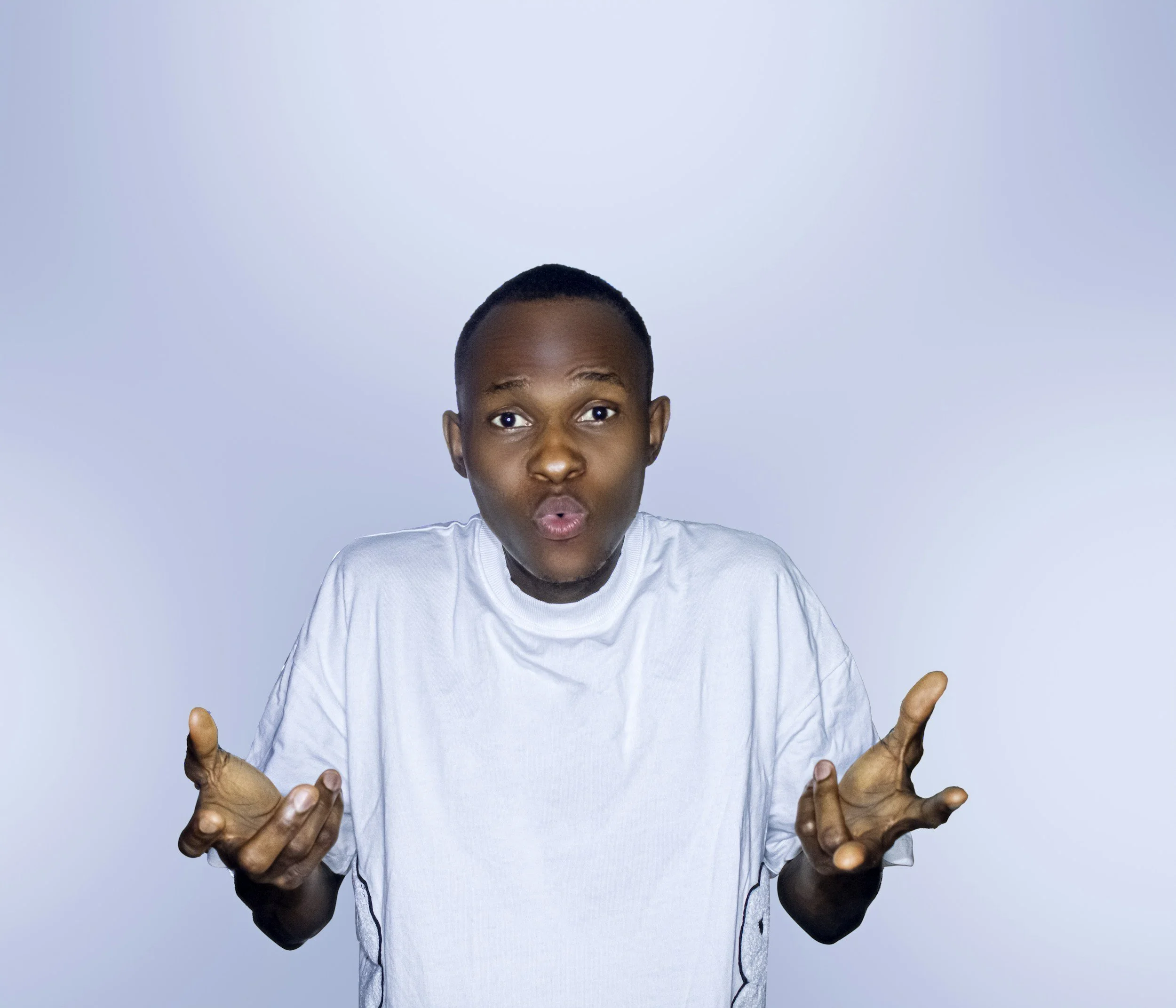 A young man with short black hair and brown skin, making an expressive face with his lips pursed and eyebrows raised, wearing a white t-shirt, against a plain light blue background.