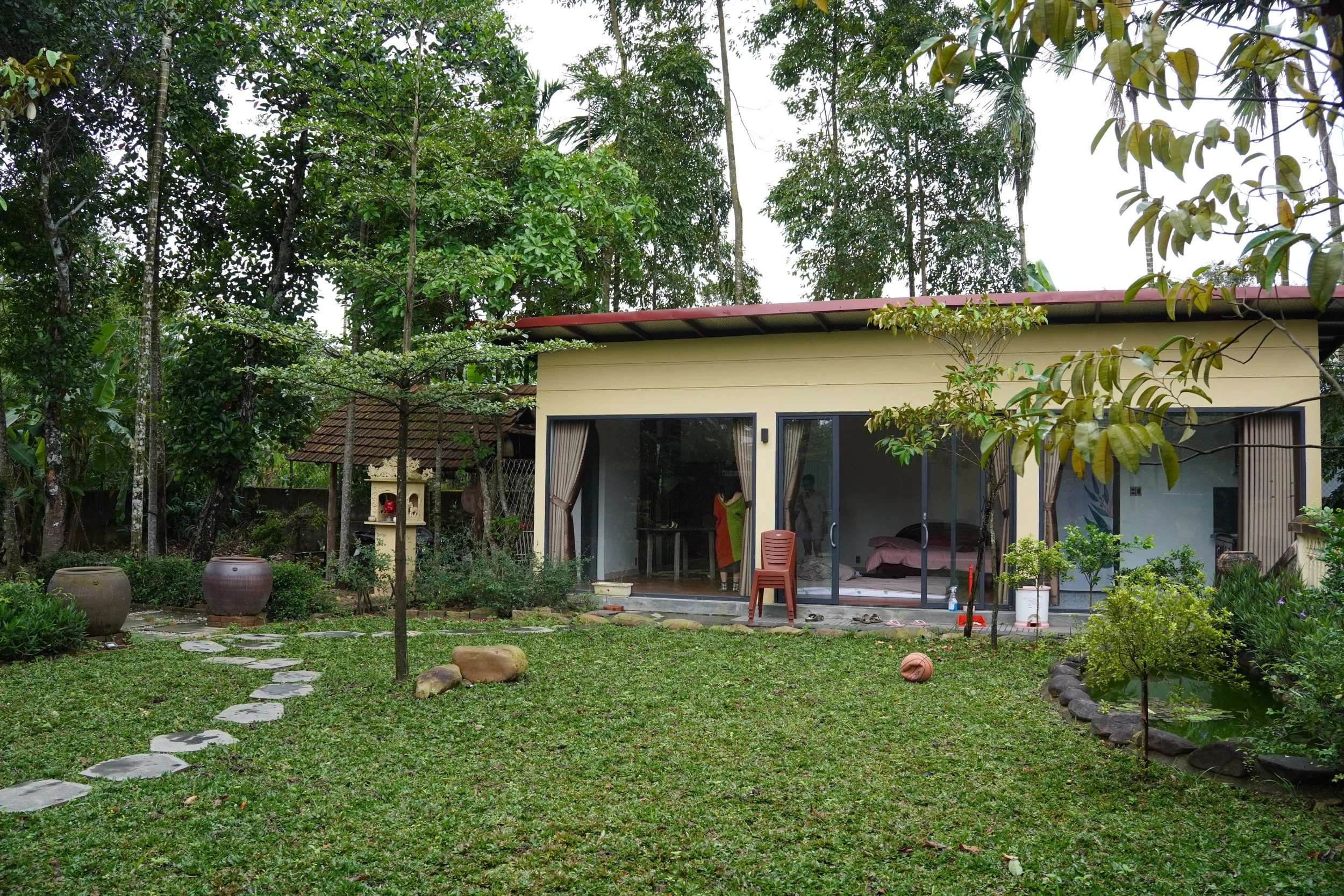 A yellow house with glass doors and curtains, surrounded by a lush green garden with trees, potted plants, and stepping stones. A person is inside the house near the glass doors and another person is visible further inside.