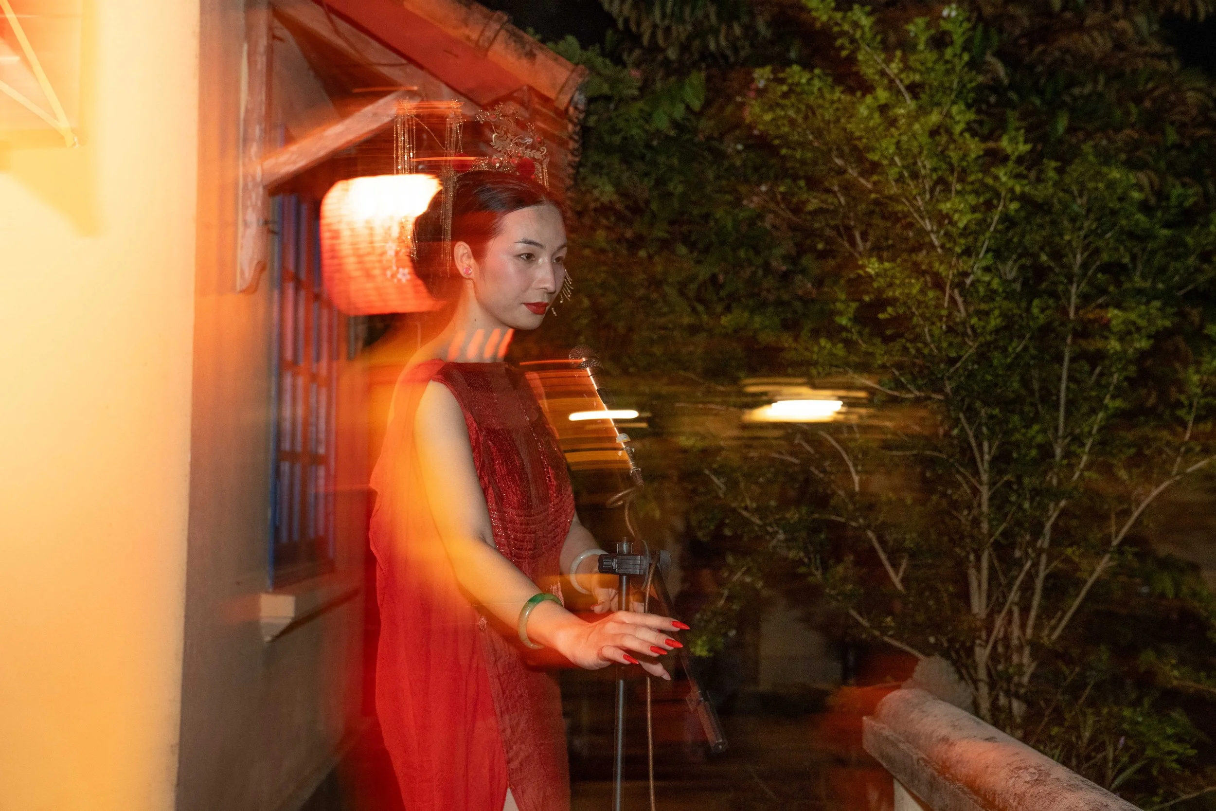 A woman dressed in a traditional red dress and green bracelet, wearing a tiara, stands on a balcony at night with a camera tripod, with trees in the background. The image has a motion blur effect.