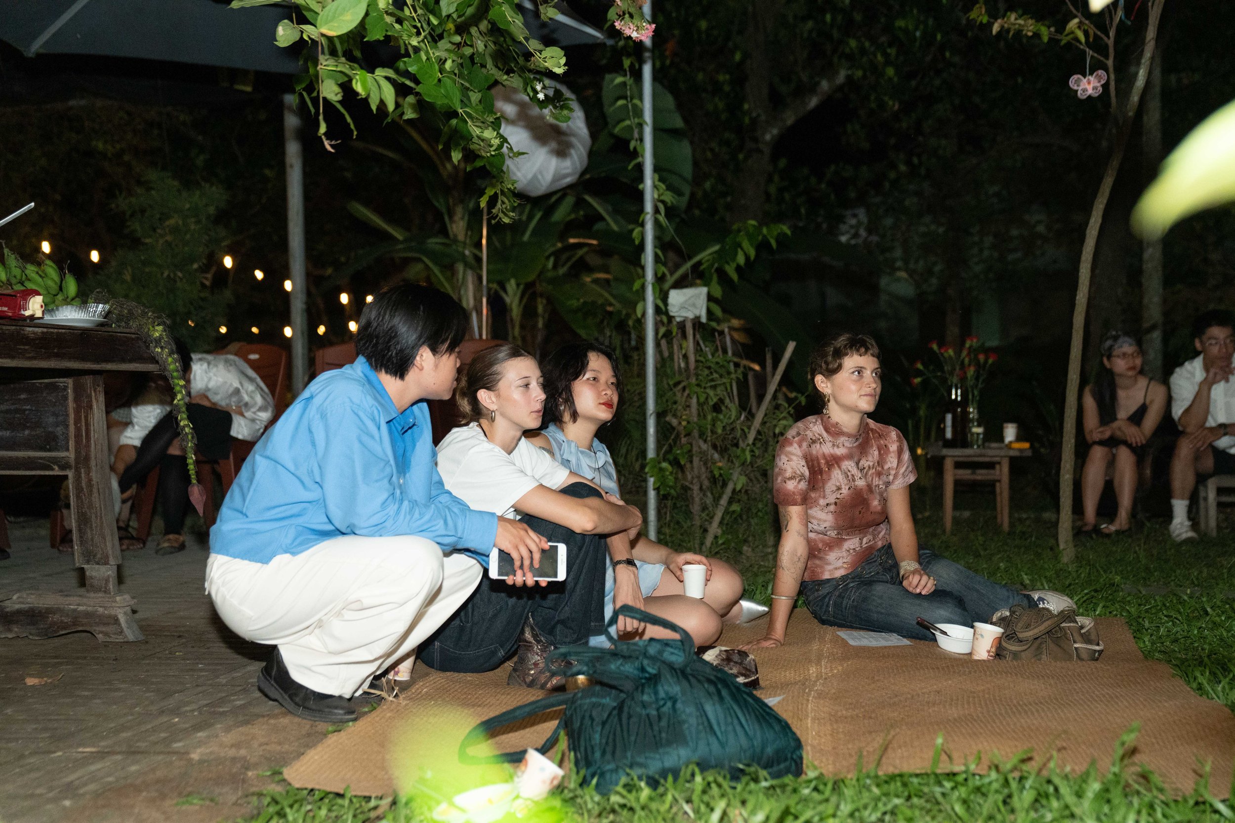 Group of people sitting on a mat outdoors at night, some with drinks, in a garden setting with trees and string lights in the background.