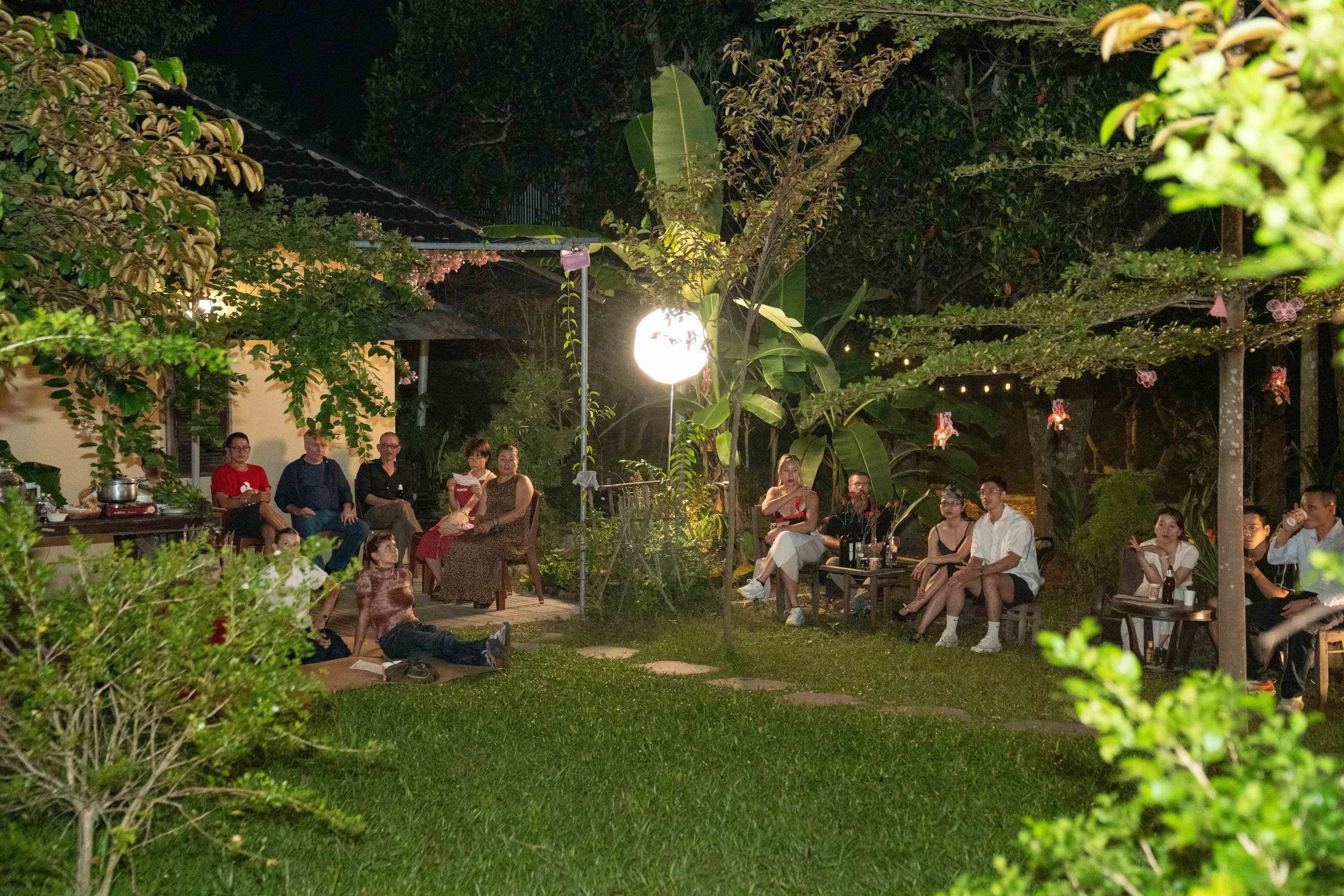 People gathered outdoors at night, sitting on chairs and benches in a garden, illuminated by a large bright light, surrounded by green trees and plants.