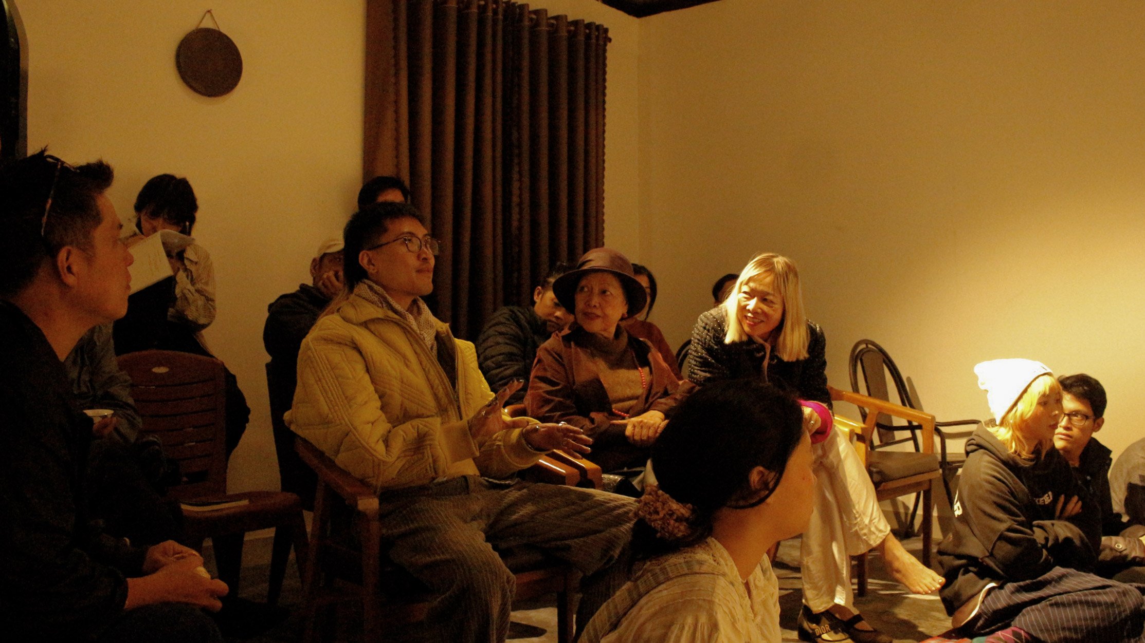 A group of people sitting and talking in a warmly lit room, with some seated on chairs and others on the floor, engaged in conversation.