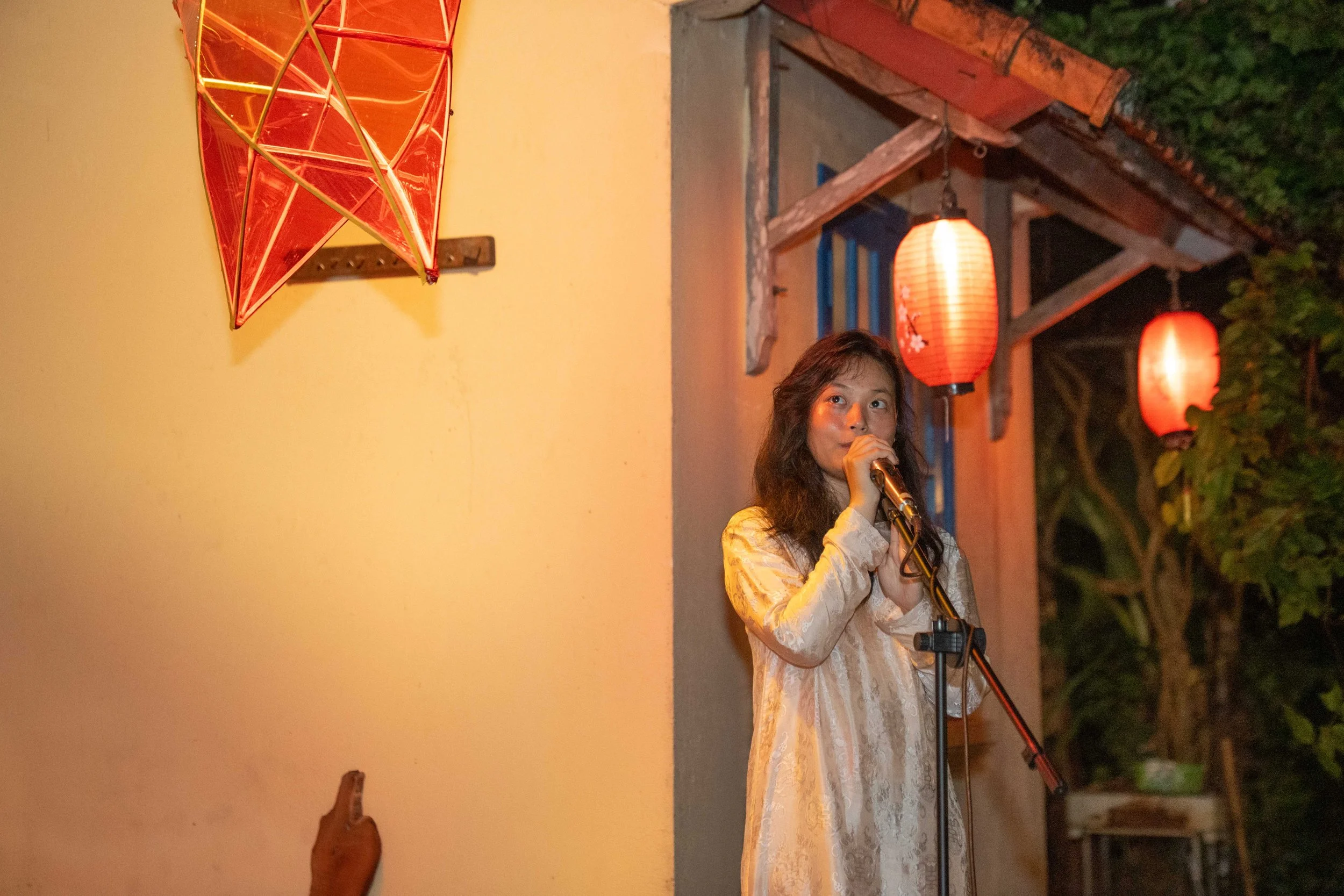 A woman in a beige dress stands on a stage, holding a microphone and looking to her right. The stage is decorated with red lanterns and a red kite on the wall in the background. It is night, and there is greenery visible to the right.