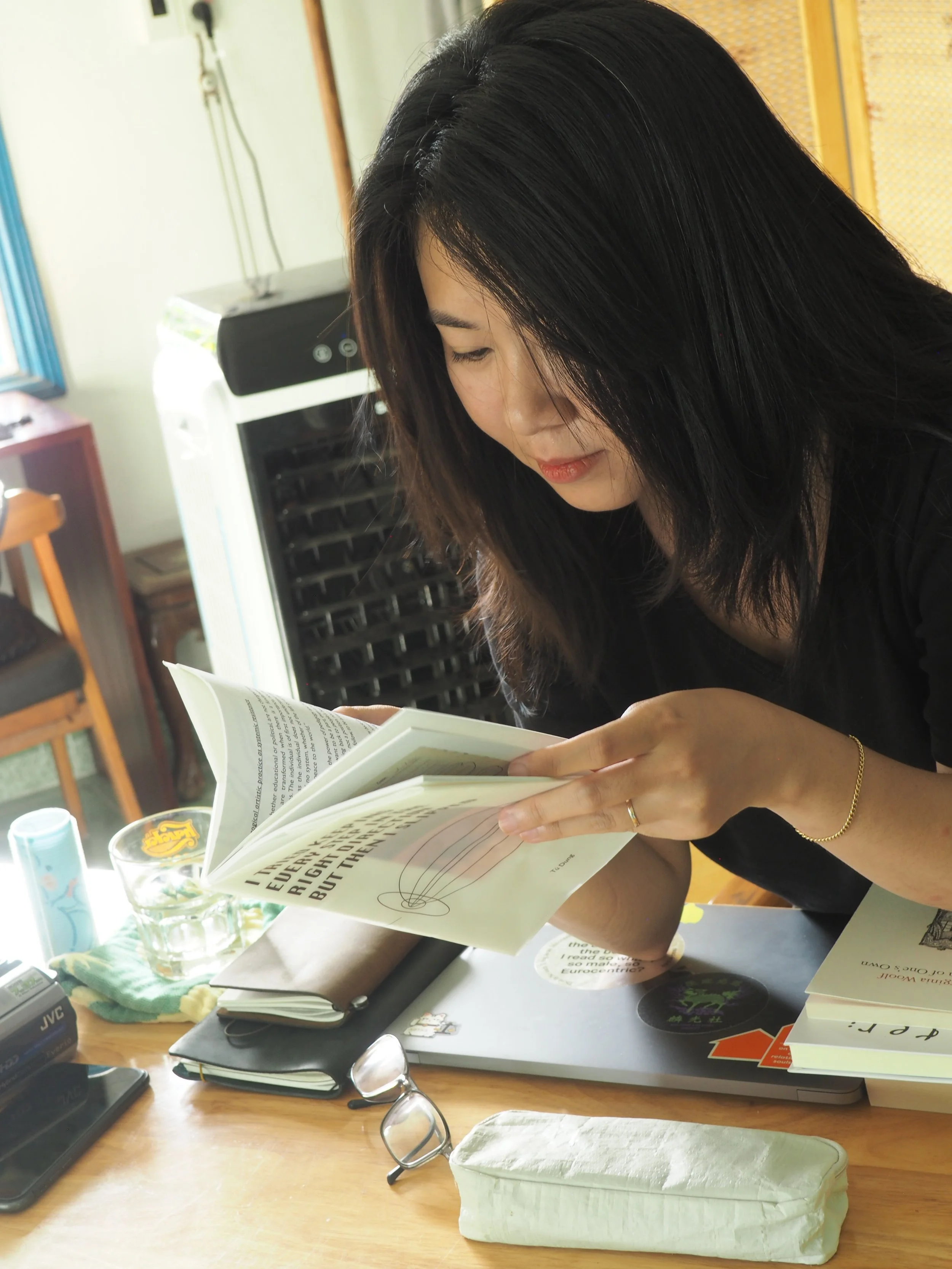 A woman with black hair is reading a book at a cluttered wooden desk. The desk has glasses, a notebook, a pen, and several other items on it. Behind her is a small white appliance and a yellow partition.