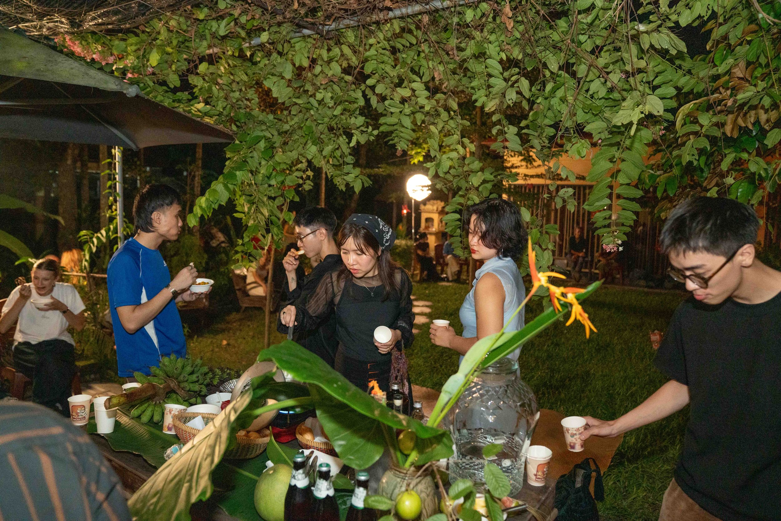 People gathered outdoors at night, standing around a table with drinks and tropical fruit, under leafy trees, enjoying a social event.