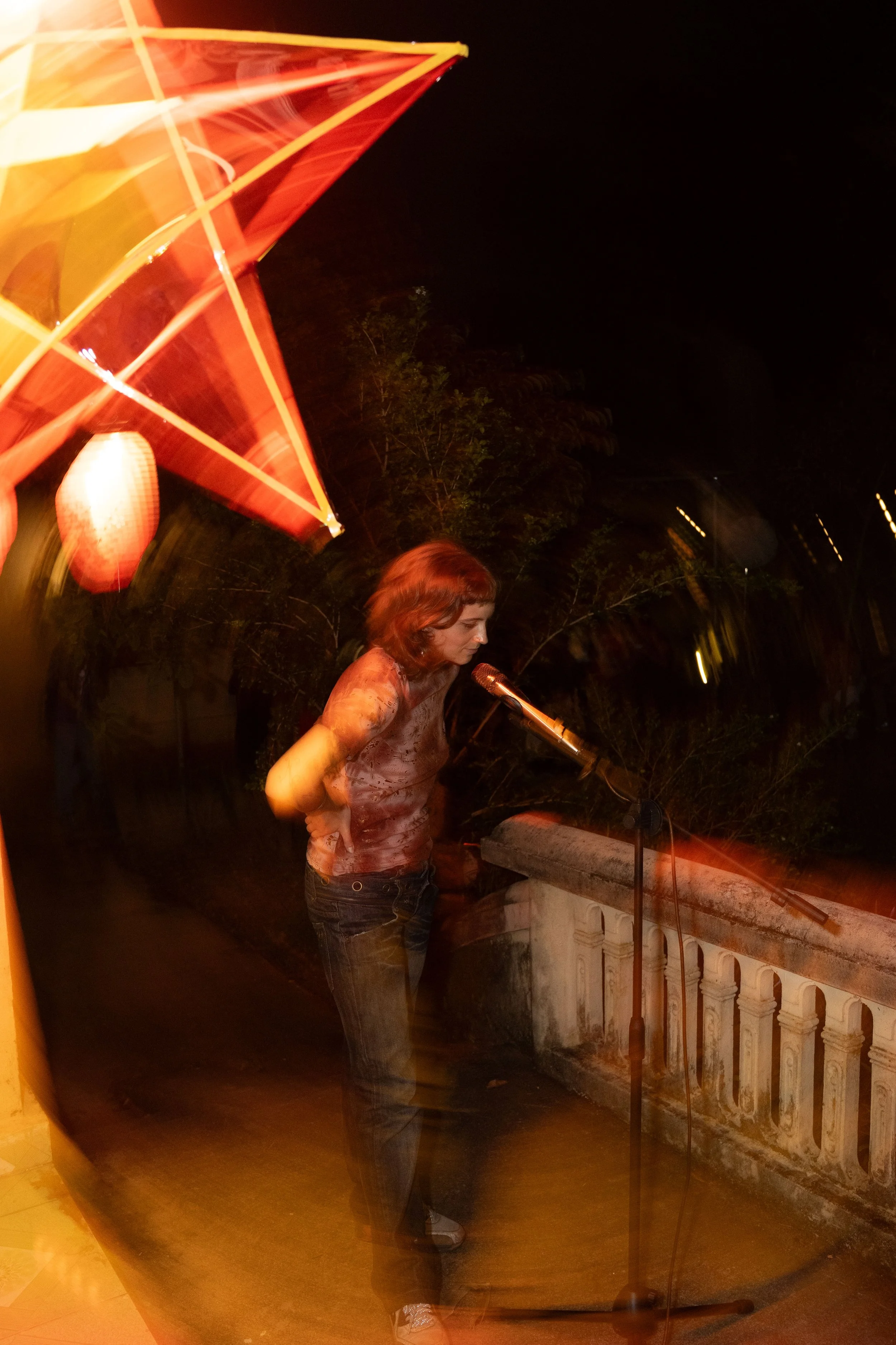 A woman with short brown hair standing on a balcony at night, reading or speaking into a microphone, with a red and yellow umbrella overhead and trees in the background.