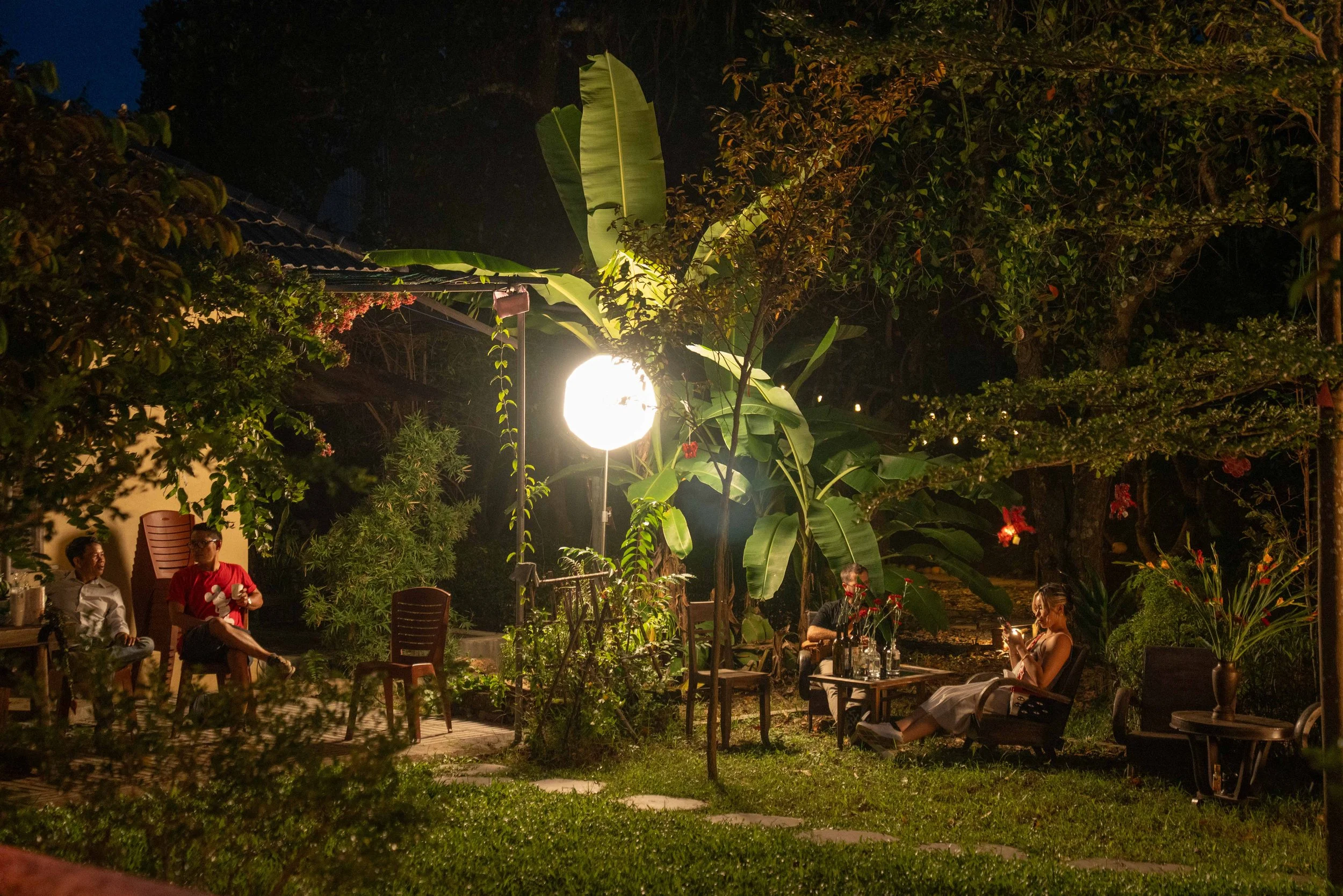 People sitting and talking in an outdoor garden at night, illuminated by a bright artificial light, surrounded by lush green plants and trees.