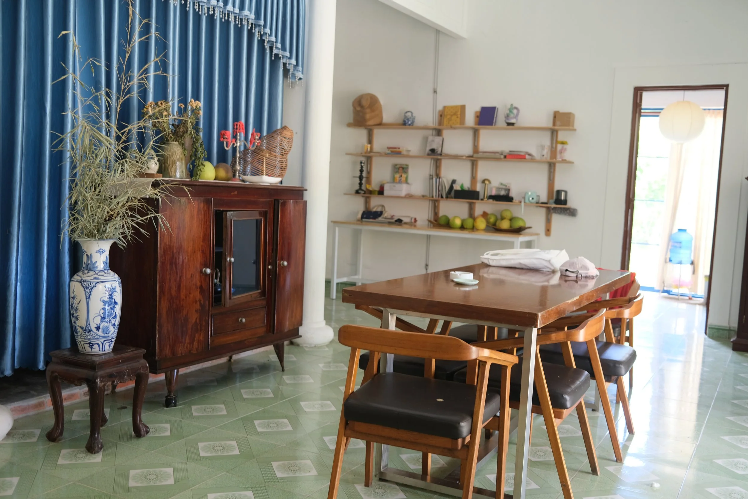 A dining room with a wooden table and six chairs, a cabinet with vases and decorative items, a blue curtain, and a wall shelf with books and green fruit. A doorway leads to a balcony with a water dispenser outside.