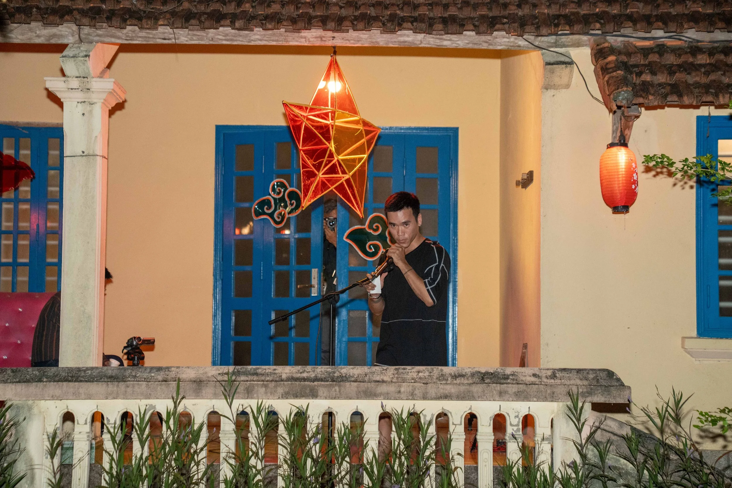 A young man is singing into a microphone on a balcony decorated with colorful lanterns and a star-shaped lantern, during an outdoor evening event.