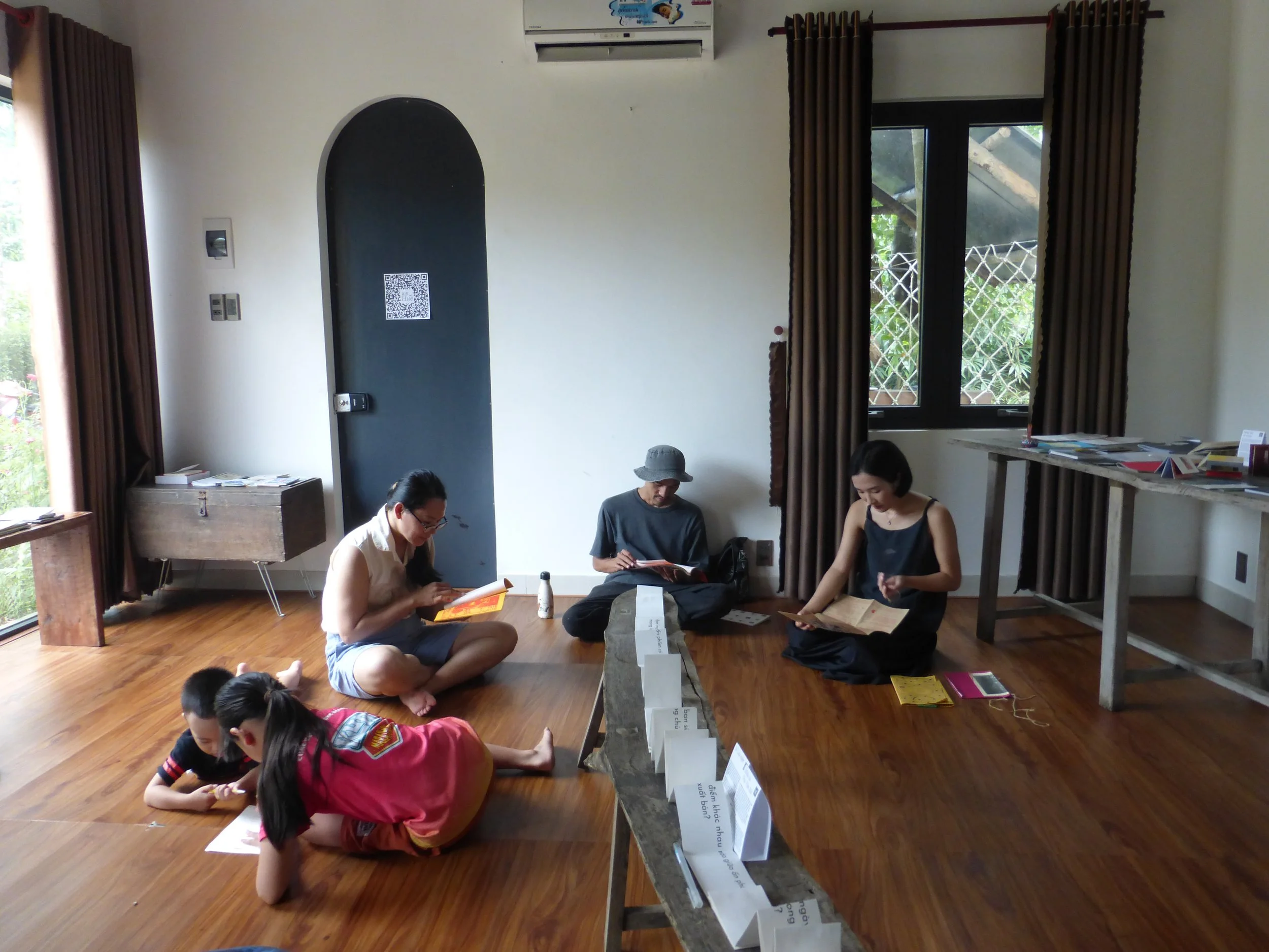 People sitting on the wooden floor reading books, two children and three adults, in a room with large windows and brown curtains.