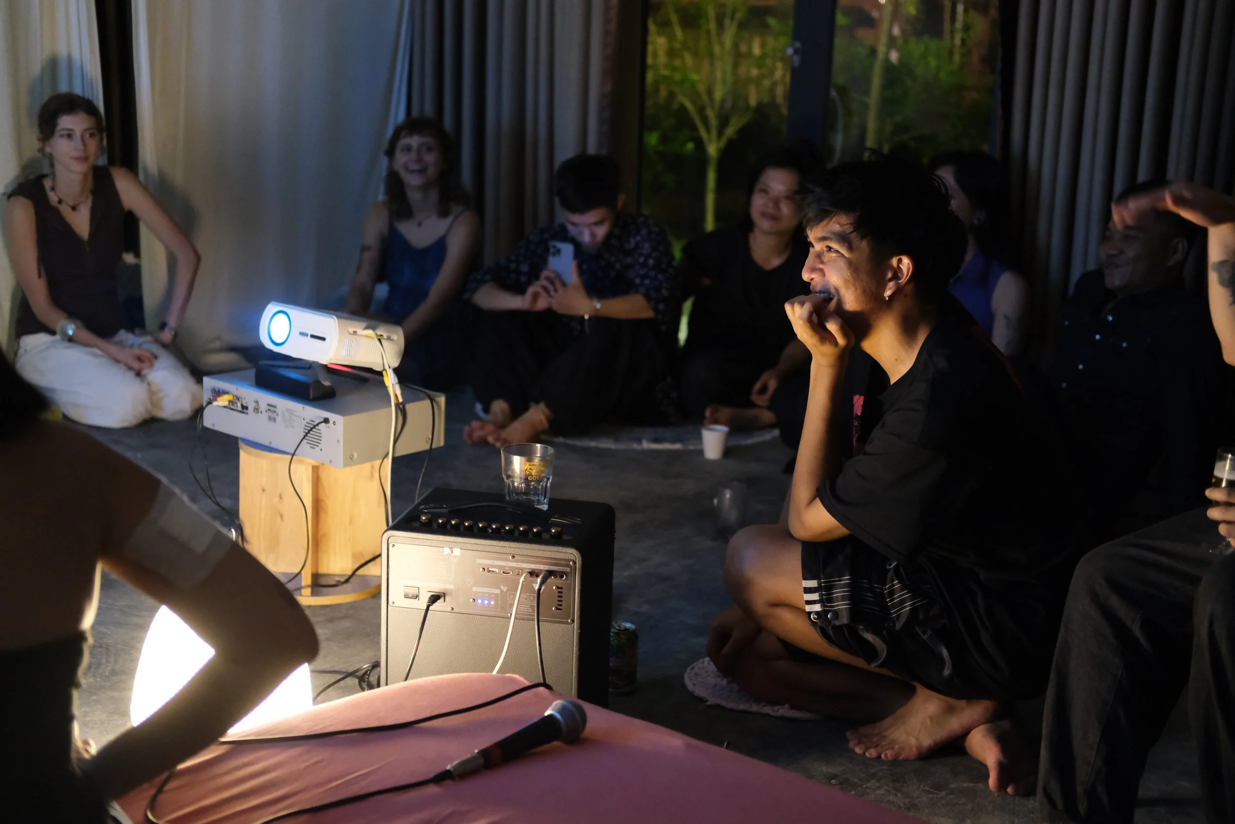 Group of people sitting on the floor, watching a projection in a dimly lit room with large windows and curtains, some smiling and one person holding a smartphone.