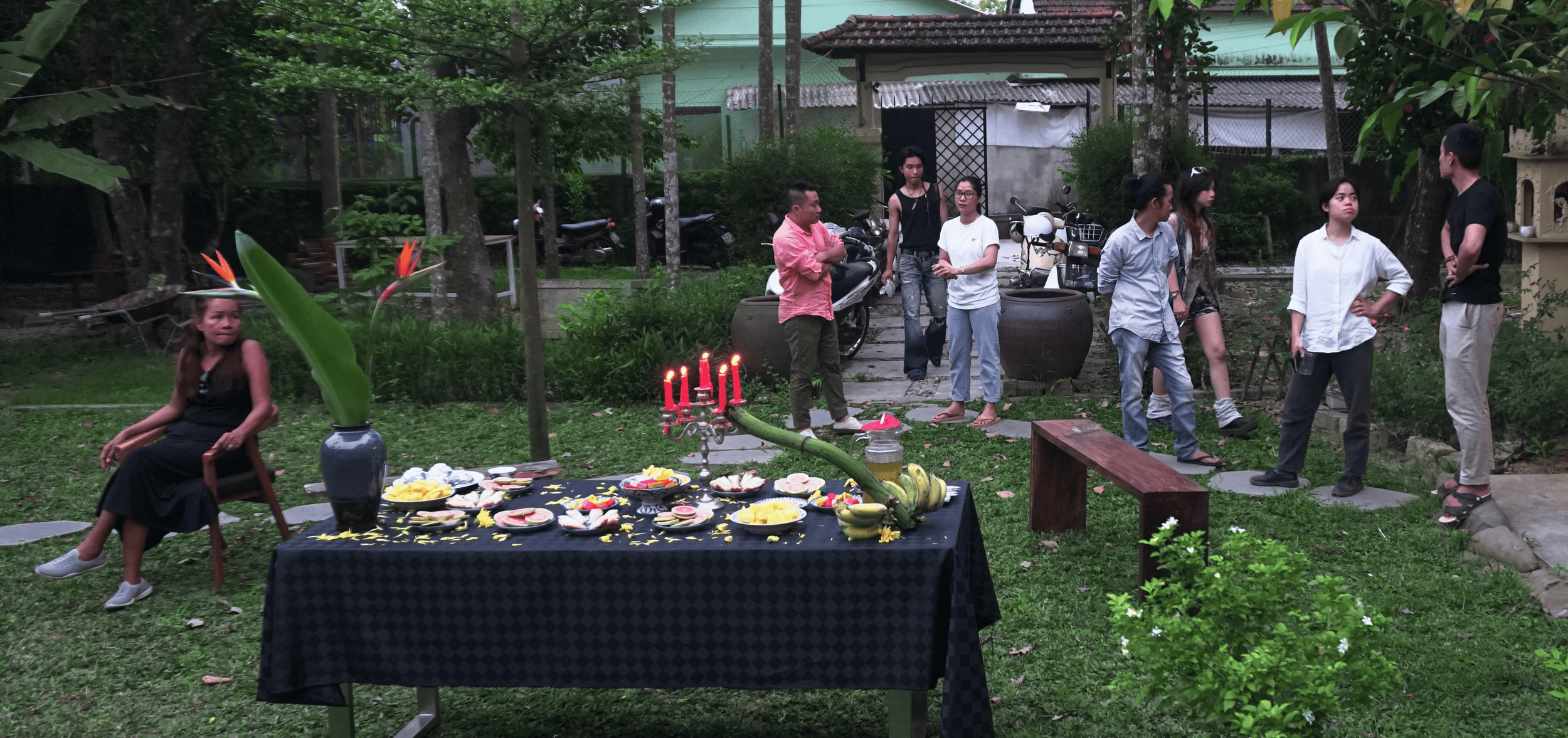 A group of people gathering outdoors in a garden for a celebration, with a table of food and a lit candelabra, surrounded by trees and plants.
