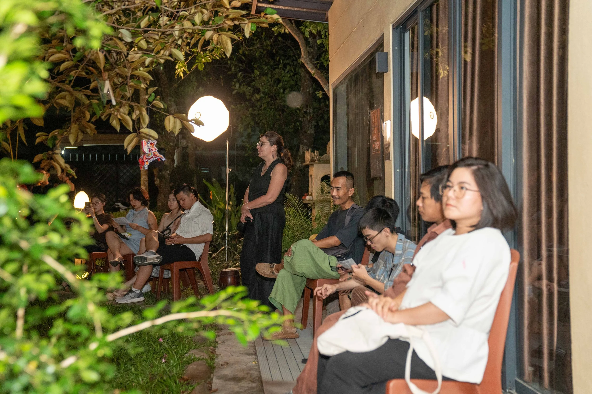 People sitting on chairs outside at night, some using their phones, with a woman standing in the background near a garden and a large light, in a social gathering setting.