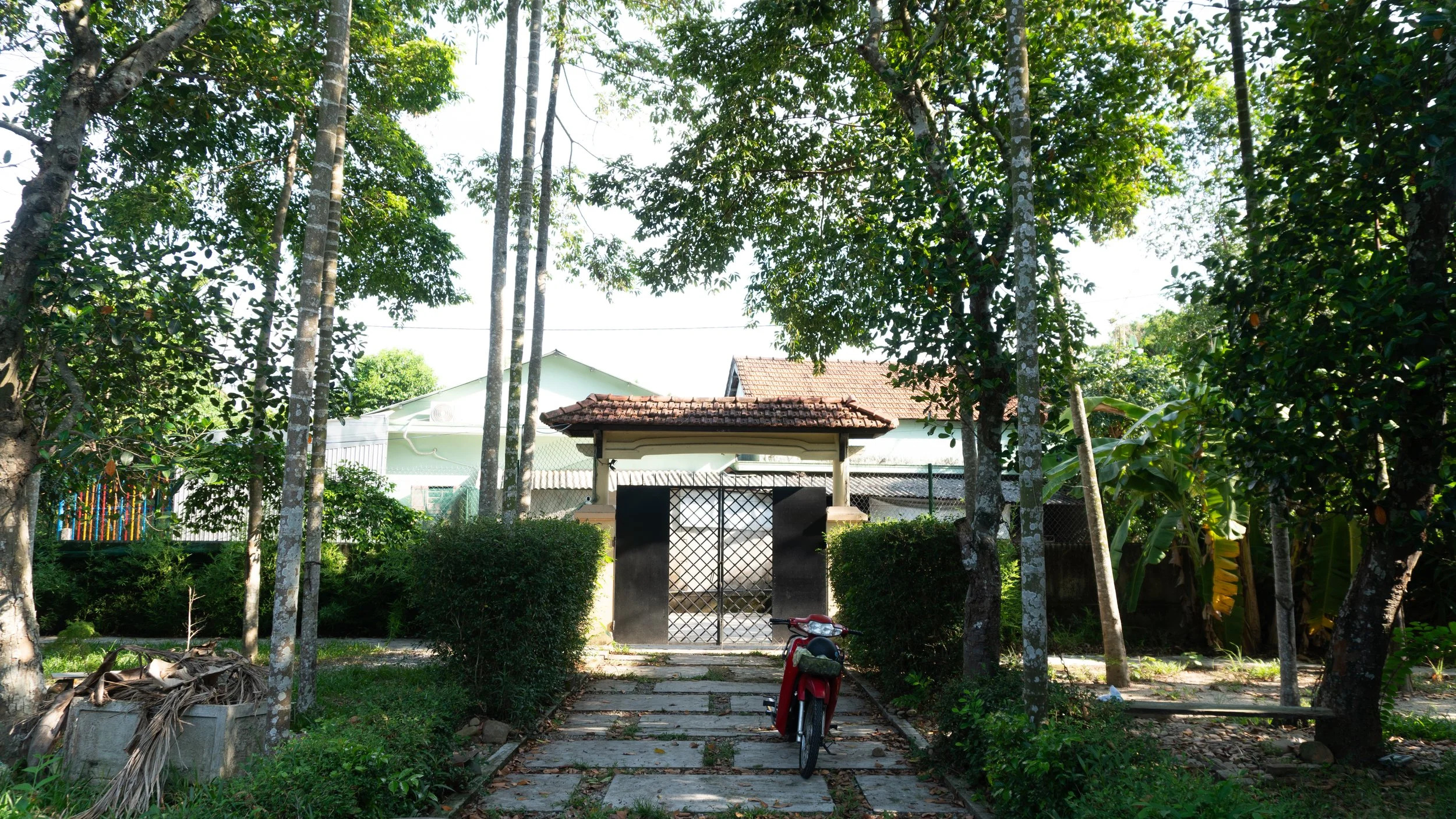 A garden with a stone pathway leading to black gate and small house with tiled roof. A red bicycle is parked on the pathway. Surrounding trees and bushes, sunlight filtering through leaves.