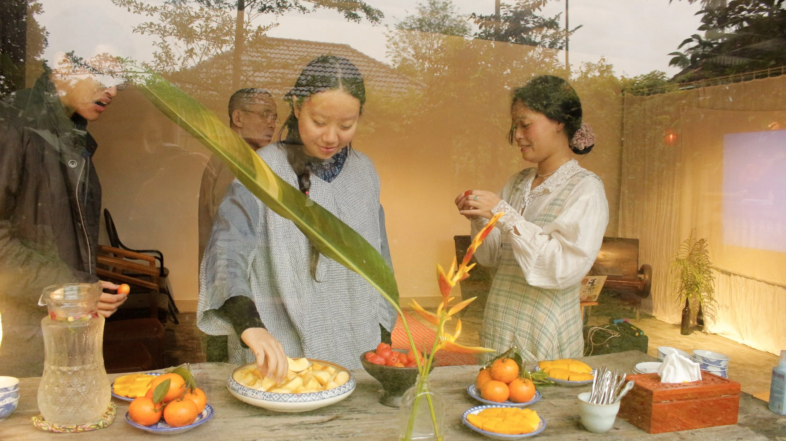 Four people are gathered around a table with bowls of sliced mango, oranges, and cherry tomatoes. One woman is holding a bowl, another is taking a piece of fruit, and a third is inspecting a small fruit in her hands. The scene is viewed through a gla