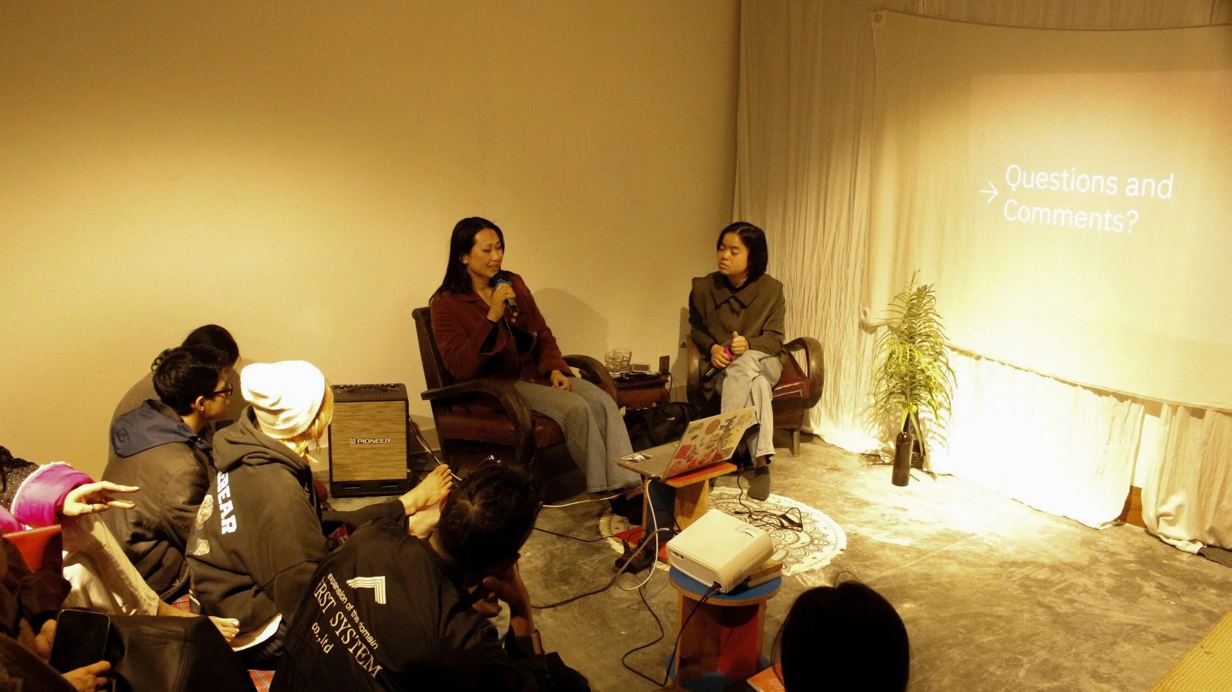 A woman speaking into a microphone while sitting on a chair and a woman sitting next to her on a chair listening. Several people are seated in front, watching and listening in a room with a large projector screen displaying the text 'Questions and Co