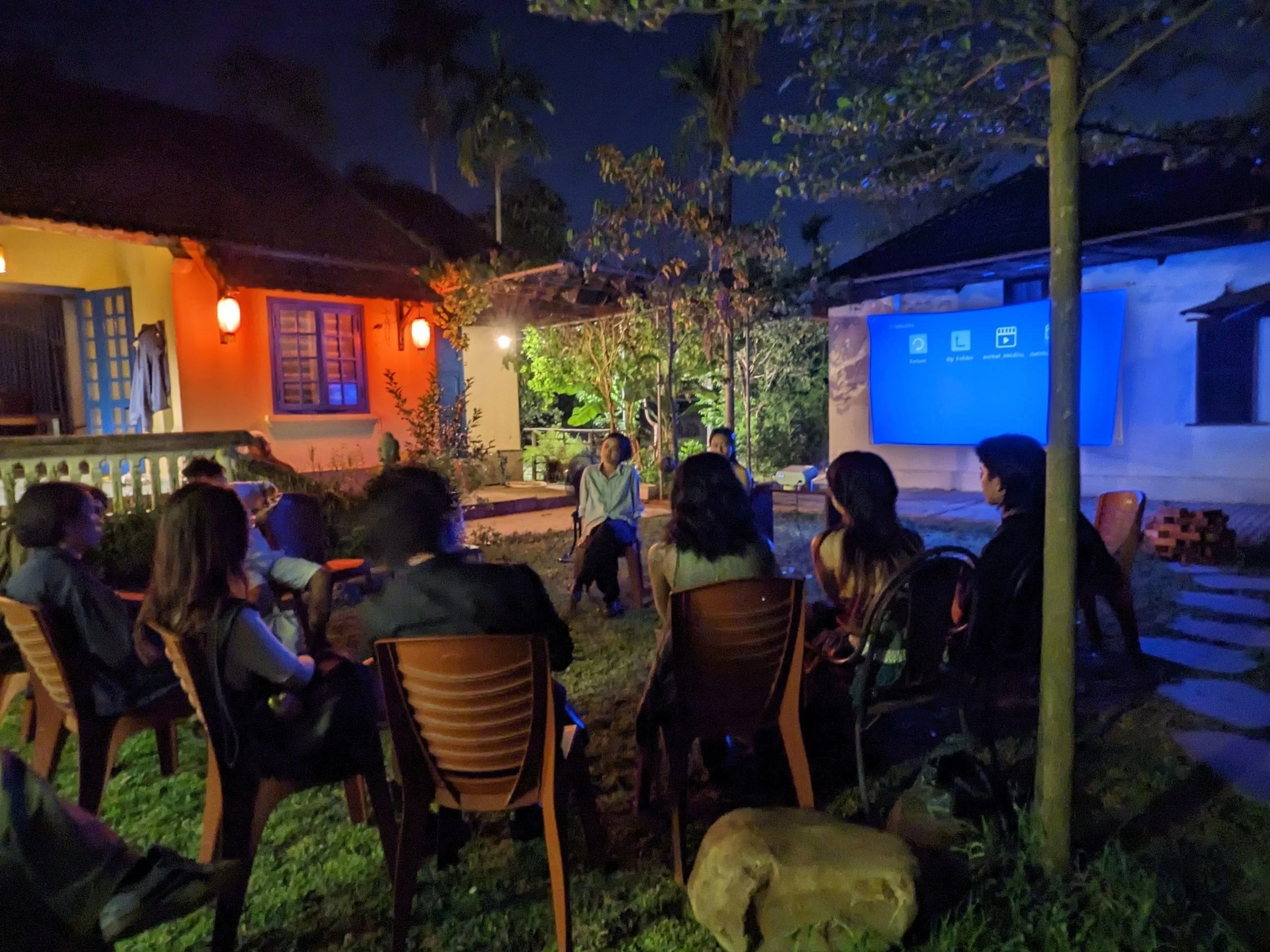 People sitting outdoors in chairs watching a large screen at night in a backyard with trees and houses.