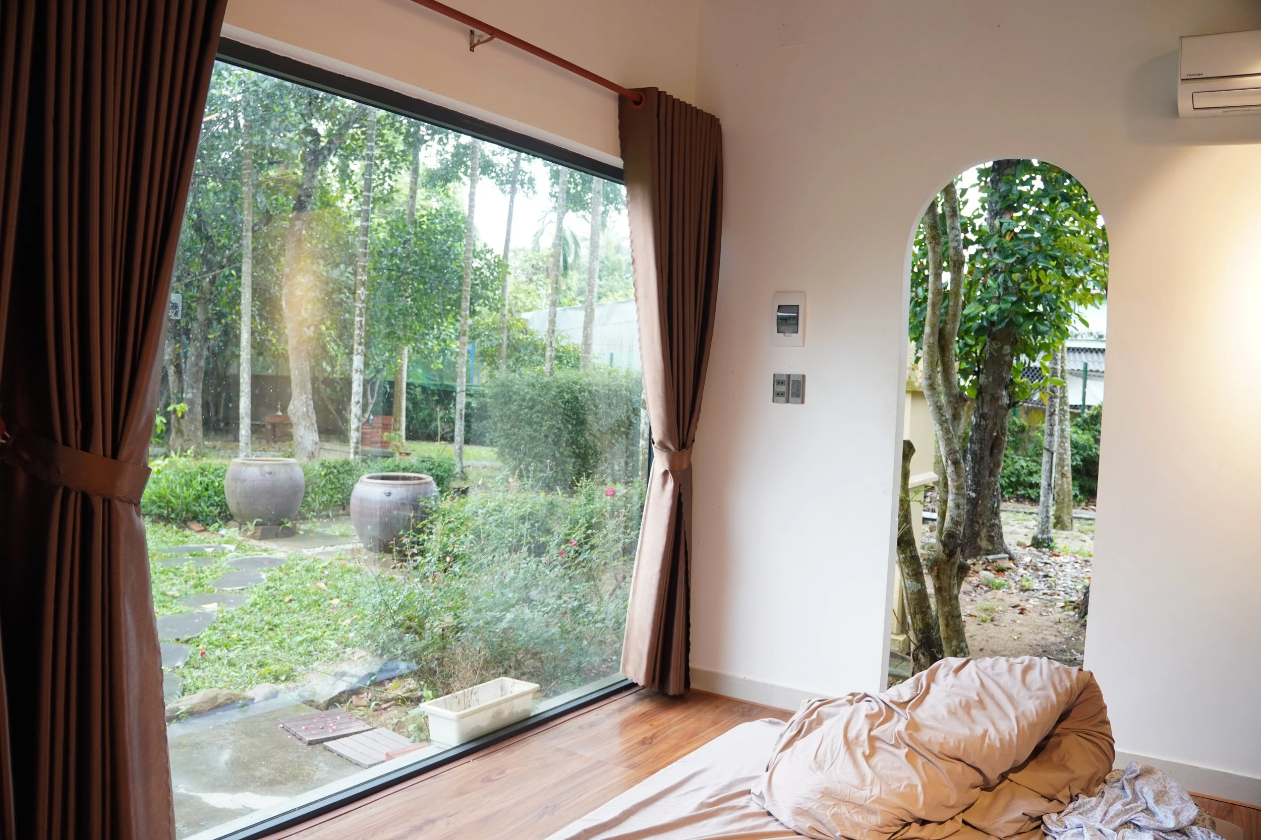 A cozy interior room with a large glass window showcasing a lush garden outside, brown curtains pulled to the sides, and a crumpled blanket on a bed or couch near the window.