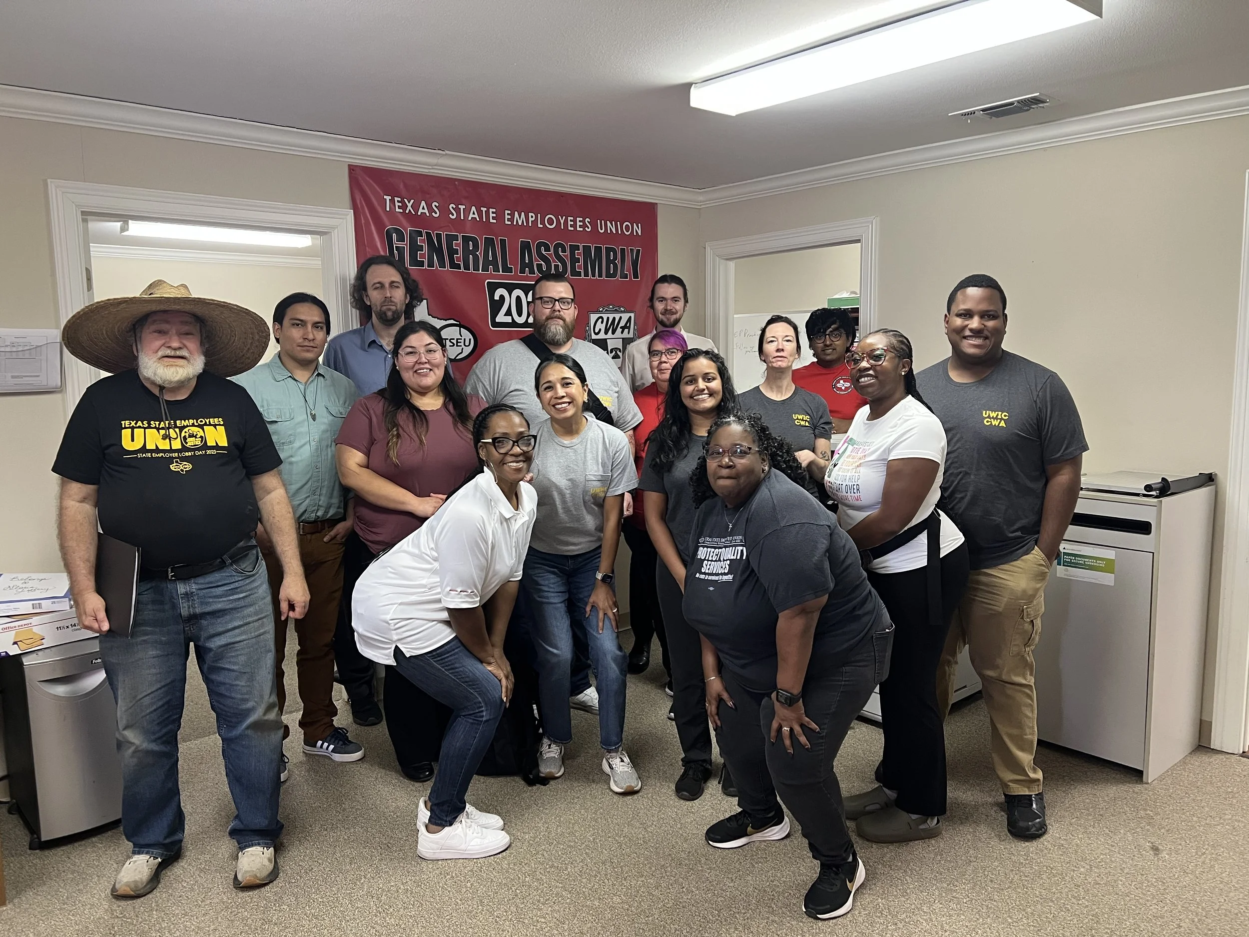 A large group of people smiling in union t-shirts in front of a red banner that reads "Texas State Employees Union General Assembly"